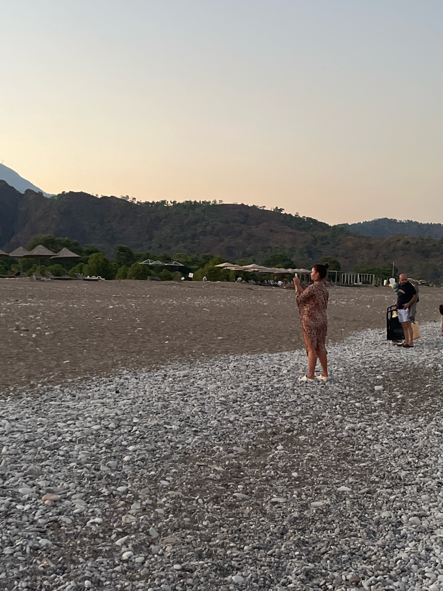 A wide-angle, eye-level, outdoor photograph captures a pebble beach in Kemer, Turkey, during the soft light of either sunrise or sunset. The sky is a pale, gradient hue, transitioning from a light yellow near the horizon to a soft blue overhead. In the distance, rolling hills covered in sparse vegetation rise, with a prominent mountain peak visible on the far left. Along the shoreline, beach umbrellas are sparsely dotted across a sandy area.
In the foreground, the beach is covered in a dense layer of smooth, grey pebbles. Three people are present. On the left, a woman stands facing away from the camera, holding a phone in her right hand as if taking a photo. She is wearing a short, patterned dress and white slides. To her right, a man stands holding a wheeled suitcase, dressed in a dark t-shirt and light shorts. Further to the right, partially out of frame, is the shoulder of another person. The overall atmosphere is serene and relaxed, suggesting a tranquil moment on the coast.
