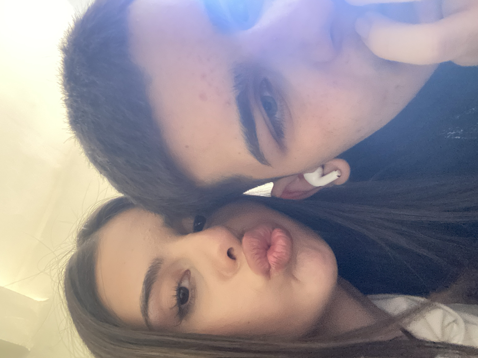 Two individuals are closely positioned, likely in a selfie-style photo. The person at the top has short, dark hair and is wearing a white earbud in their right ear. The person at the bottom has long, dark hair and is pouting with their lips. The environment is poorly lit and indistinct. The location of the city of Sofia, Bulgaria cannot be determined based on the image.