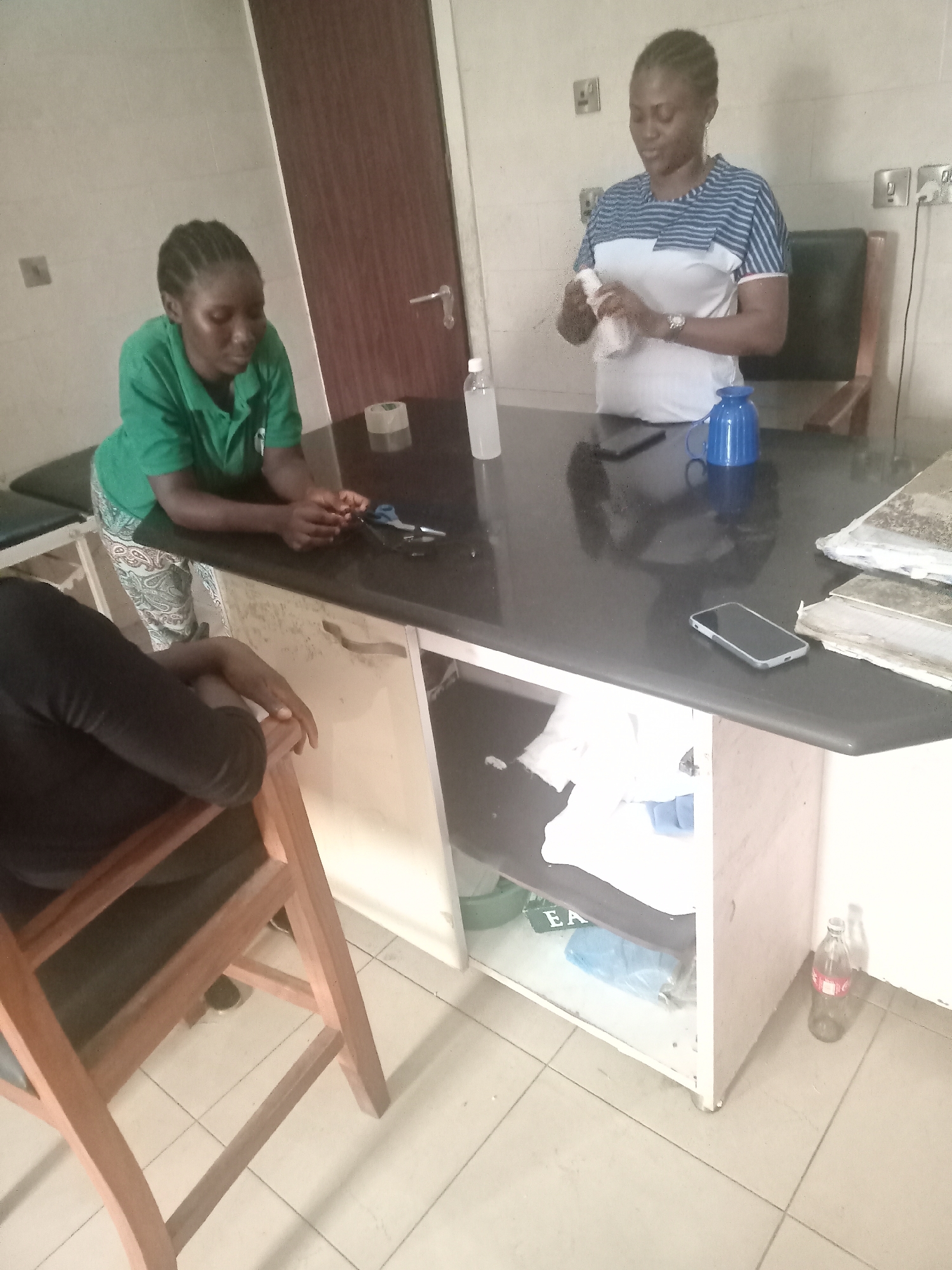 The photograph, taken in what appears to be an office in Gwarinpa, Nigeria, shows two women standing behind a large desk. One woman is wearing a green t-shirt and patterned trousers, leaning on the desk, with scissors and tape measure in front of her. The other woman, in a blue and white striped shirt, is holding a bottle of hand sanitizer. The desk is black, with a bottle of sanitizer, a blue mug, a cellphone, and a pile of papers on top. Underneath the desk, the open cabinet reveals assorted items. A person in a black shirt sits on a wooden chair to the left of the desk. An empty coca-cola bottle stands on the floor beside the desk. The walls are light-colored and there are electrical outlets visible. A closed door is also visible.
