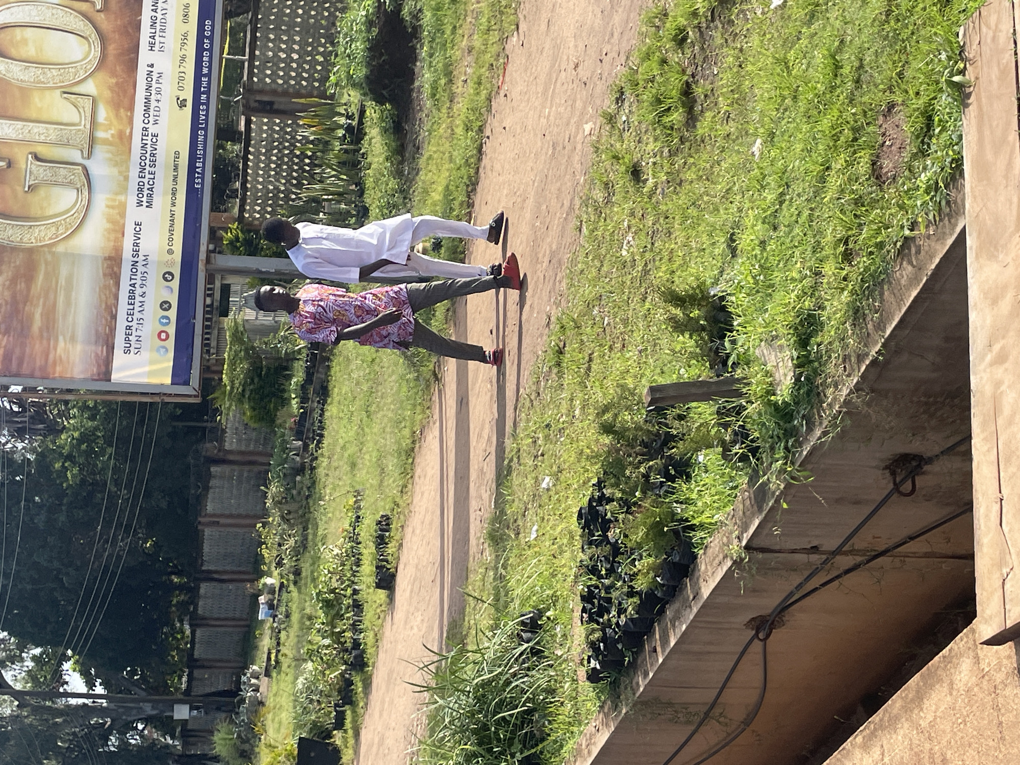 Two young men walk along a dirt path lined with numerous potted plants and a concrete drainage channel. The man on the left wears a patterned short-sleeved shirt and dark pants with red sandals. The man on the right is dressed in a white long-sleeved shirt and white pants with dark shoes. In the background, a large billboard with text and logos is visible, along with trees, a fence, and power lines. The scene appears to be outdoors, possibly in a nursery or garden area, with a distinct urban environment suggested by the billboard and infrastructure. The location is identified as Jos, Nigeria.