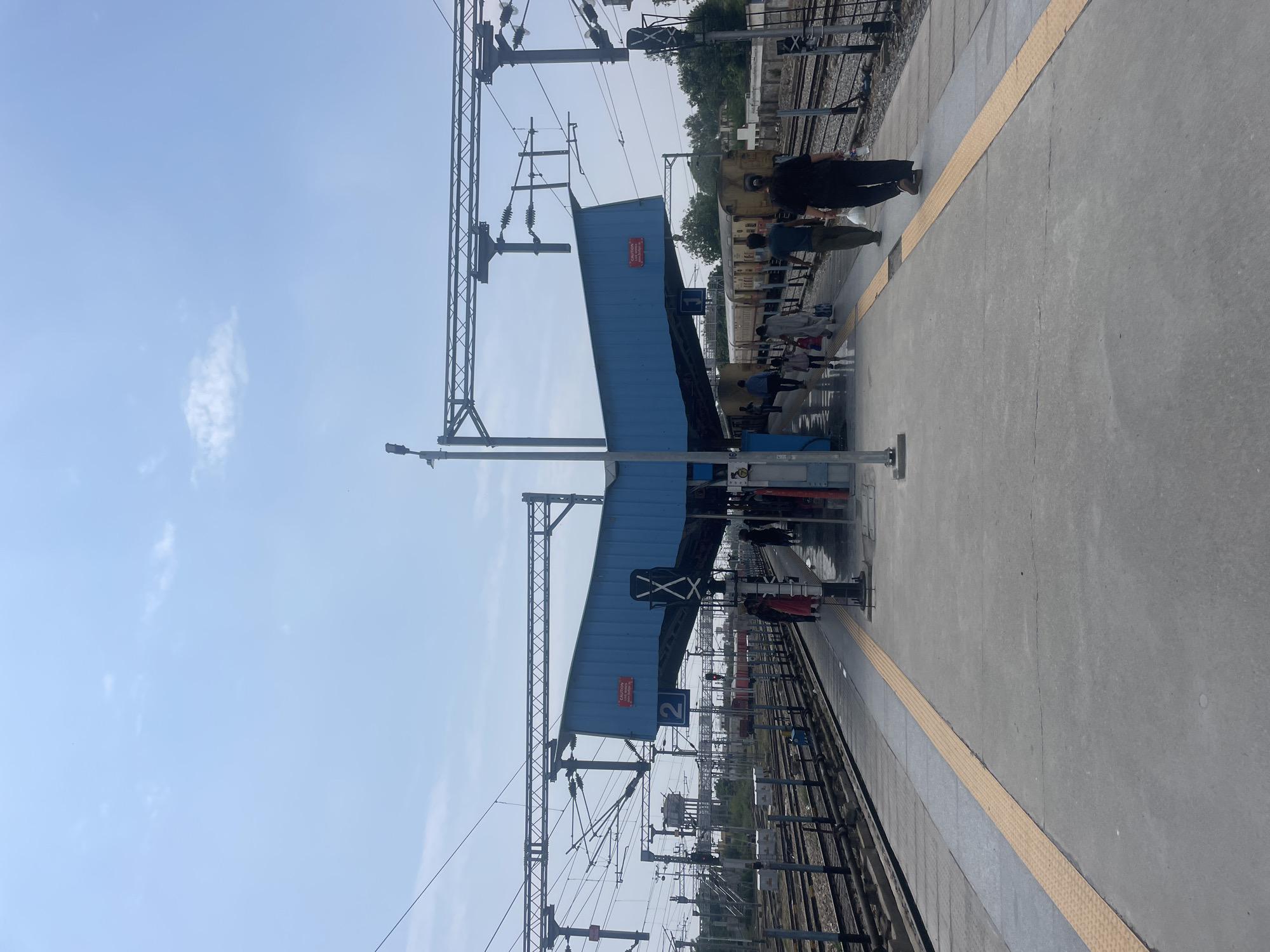 The image depicts a view of a train station platform in Pathankot, India, under a bright, partly cloudy sky. In the foreground, a concrete platform is visible, with a tactile paving strip running along its edge. Several people are walking along the platform. In the mid-ground, a blue-colored roofed structure shelters a section of the platform, with signage indicating platform number 2. Overhead, numerous electrical wires and supports for the overhead catenary system are prominent. Beyond the platform, train tracks are visible, with a train with yellow carriages present on one of the tracks. In the background, trees and buildings are visible.