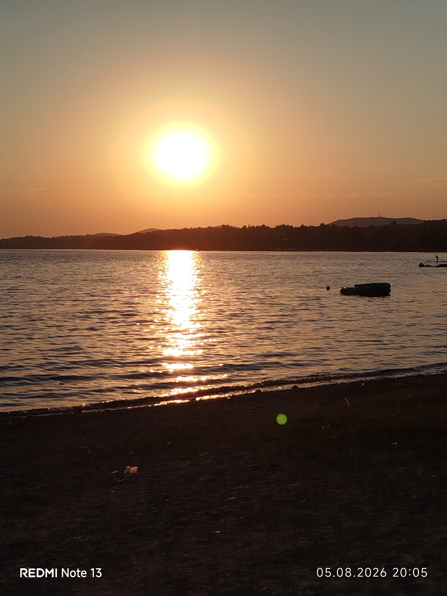 The image depicts a serene sunset over a body of water. The sun, a brilliant orb, is positioned in the upper left quadrant, casting a warm, golden glow across the sky and reflecting a shimmering path on the water's surface. The horizon is lined with a dark silhouette of trees and hills, with a faint structure resembling a radio tower visible on one of the higher points. In the foreground, a dark, sandy shore meets the water. The water itself is calm with gentle ripples. A dark, elongated object, possibly a float or debris, is visible in the middle of the water, along with a small, dark round object. Further to the right, a person is standing in the water, appearing to interact with something. The overall atmosphere is tranquil and bathed in the warm hues of sunset. The timestamp "05.08.2026 20:05" is present in the bottom right corner, indicating the date and time the photograph was taken. "REDMI Note 13" is also visible in the bottom left corner, indicating the device used to capture the image. The location is identified as Sithonia Municipal Unit, Greece.