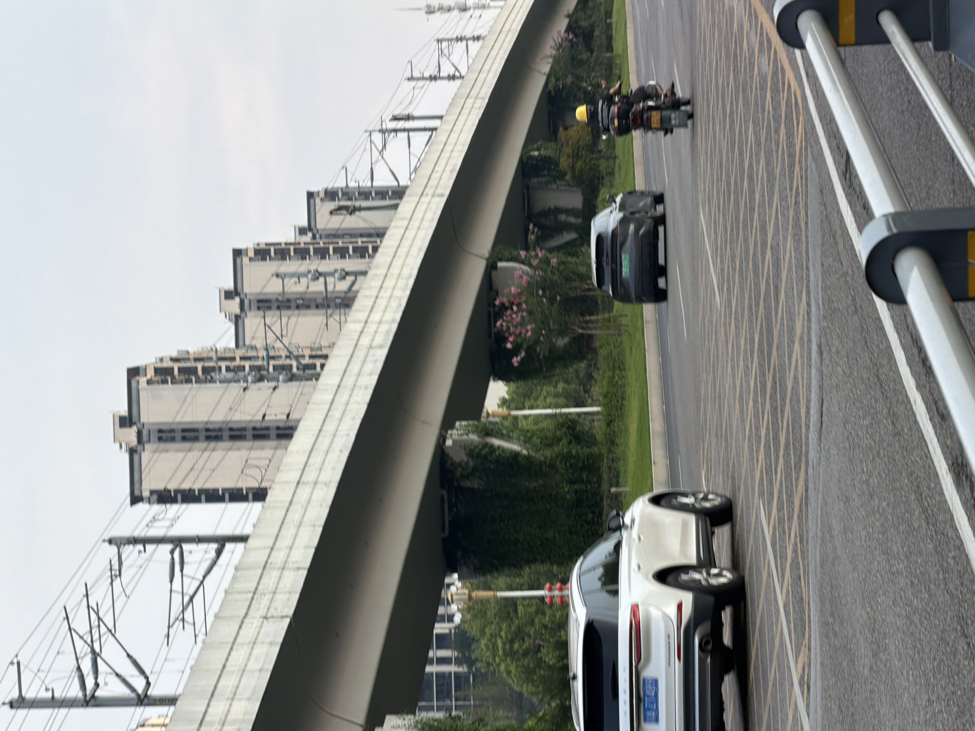 In Jiangjin, China, a white SUV is parked in the foreground on the right side of the road, facing away from the viewer. Its rear is towards the camera, and it is on a multi-lane asphalt road marked with yellow diagonal lines. A dark-colored SUV is in the middle lane, also facing away from the viewer, and a motorcycle with a person wearing a yellow helmet is in the right lane, moving away from the viewer. In the background, a concrete overpass spans across the scene, supported by pillars covered in green vegetation. Beyond the overpass, several multi-story buildings are visible against a hazy sky. Power lines and their supporting structures are prominent above and around the overpass. The scene appears to be a city street during daylight.