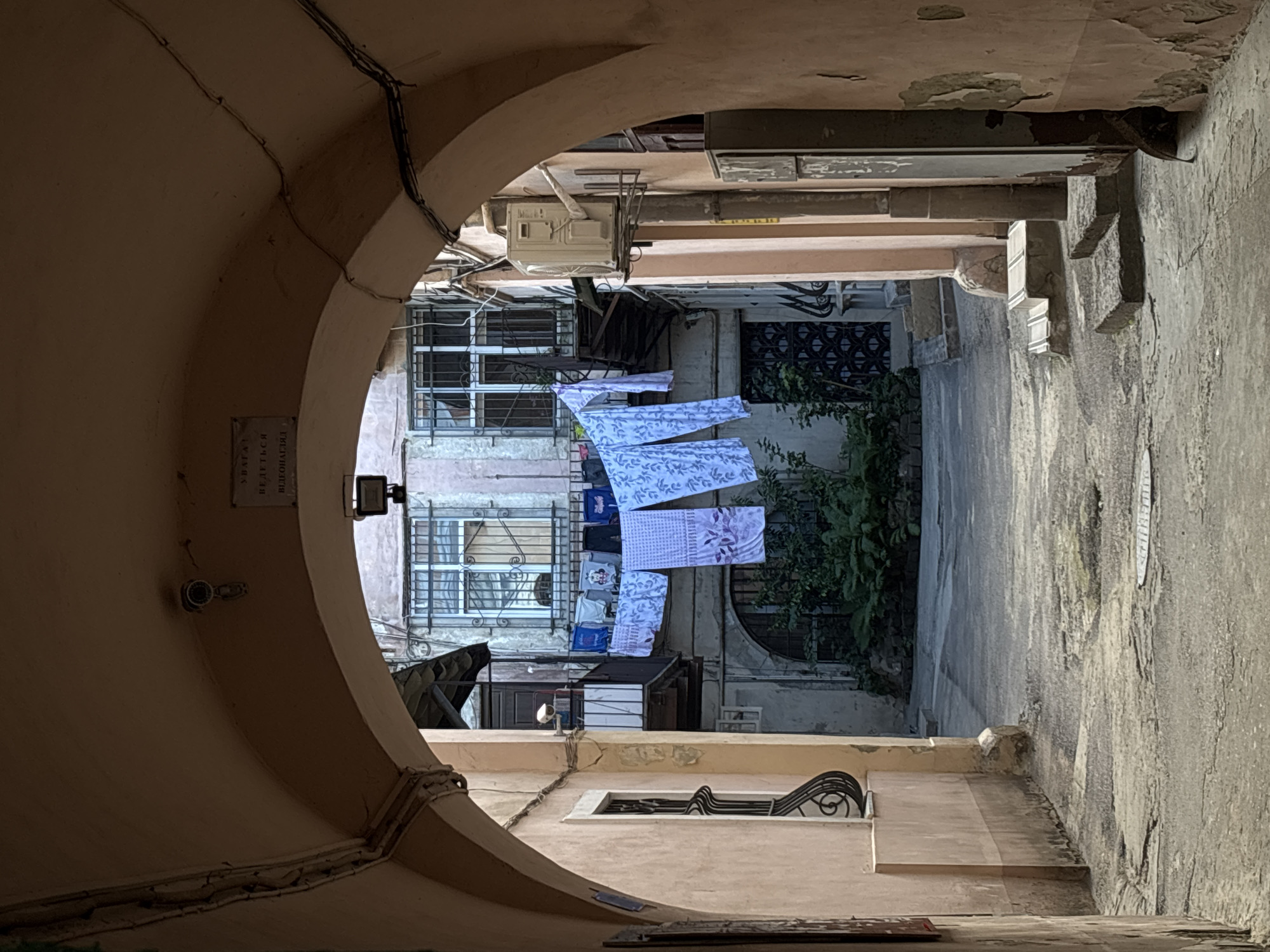 Captured from within a curved archway, the image frames a narrow courtyard scene in Lviv, Ukraine. Laundry hangs between buildings. Walls are plastered and weathered. Windows are barred. The courtyard floor is concrete. Electrical wiring is visible along the archway. A surveillance camera is mounted on the archway wall.