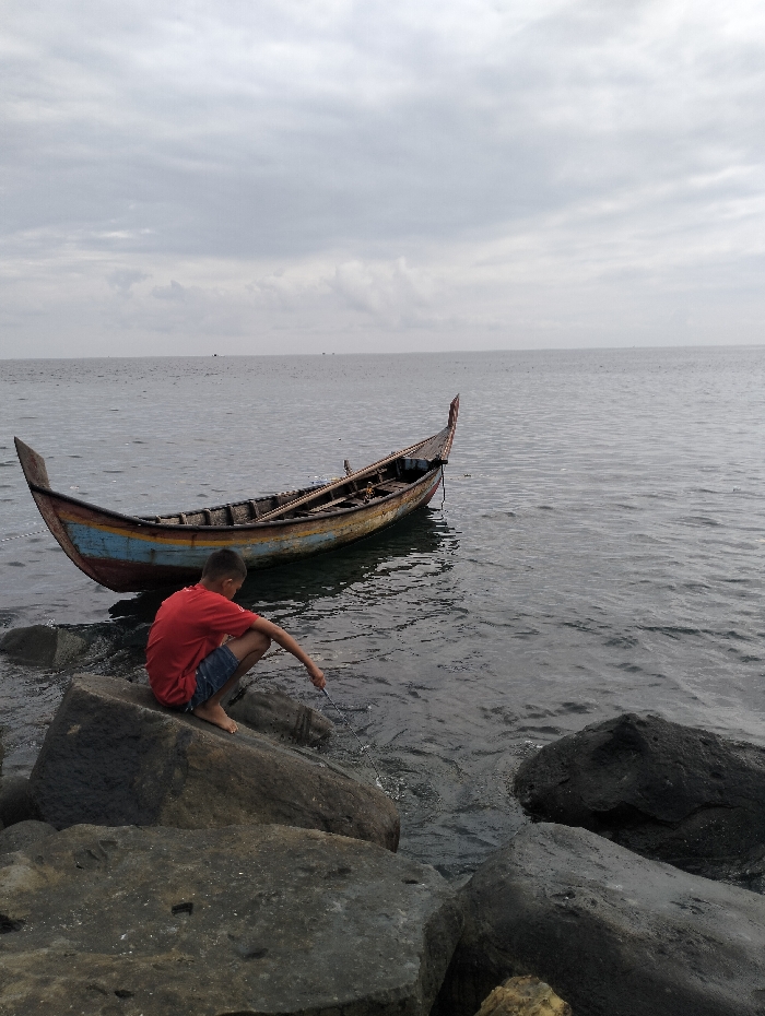 A young boy in a red shirt and blue shorts sits on dark rocks at the edge of the water, possibly fishing with a stick. An old, colorful wooden boat rests on the water's surface nearby. The sea is calm, stretching to the horizon under a cloudy, overcast sky. The scene evokes a sense of quiet reflection and a connection with nature.