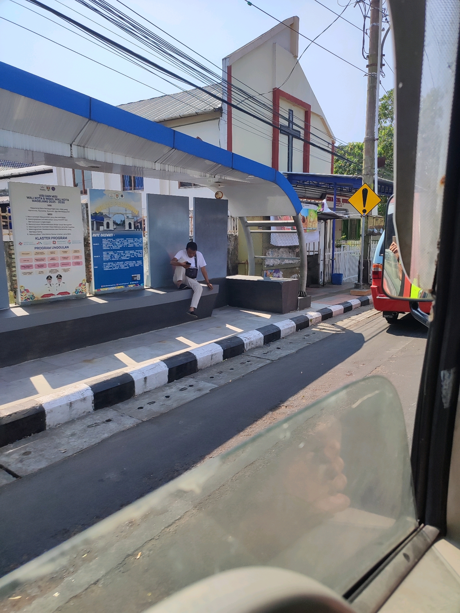 The image captures a street scene in Magelang, Indonesia, likely near a bus stop or a public facility. A man in a white shirt and light-colored pants is seated on a concrete bench, looking at his phone. He has a black bag slung across his body. Behind him, a modern building with a prominent red cross on its facade suggests a religious institution, possibly a church. A blue awning extends over a waiting area. Several informational posters are displayed on panels. The street in the foreground has a distinctive black and white striped curb. A red vehicle, possibly a bus or van, is visible in the right side of the frame, with reflections on its windows. The scene is bathed in bright sunlight, casting shadows. Overhead, a complex network of electrical wires crisscrosses the sky. A yellow triangular warning sign with an exclamation mark is also present.