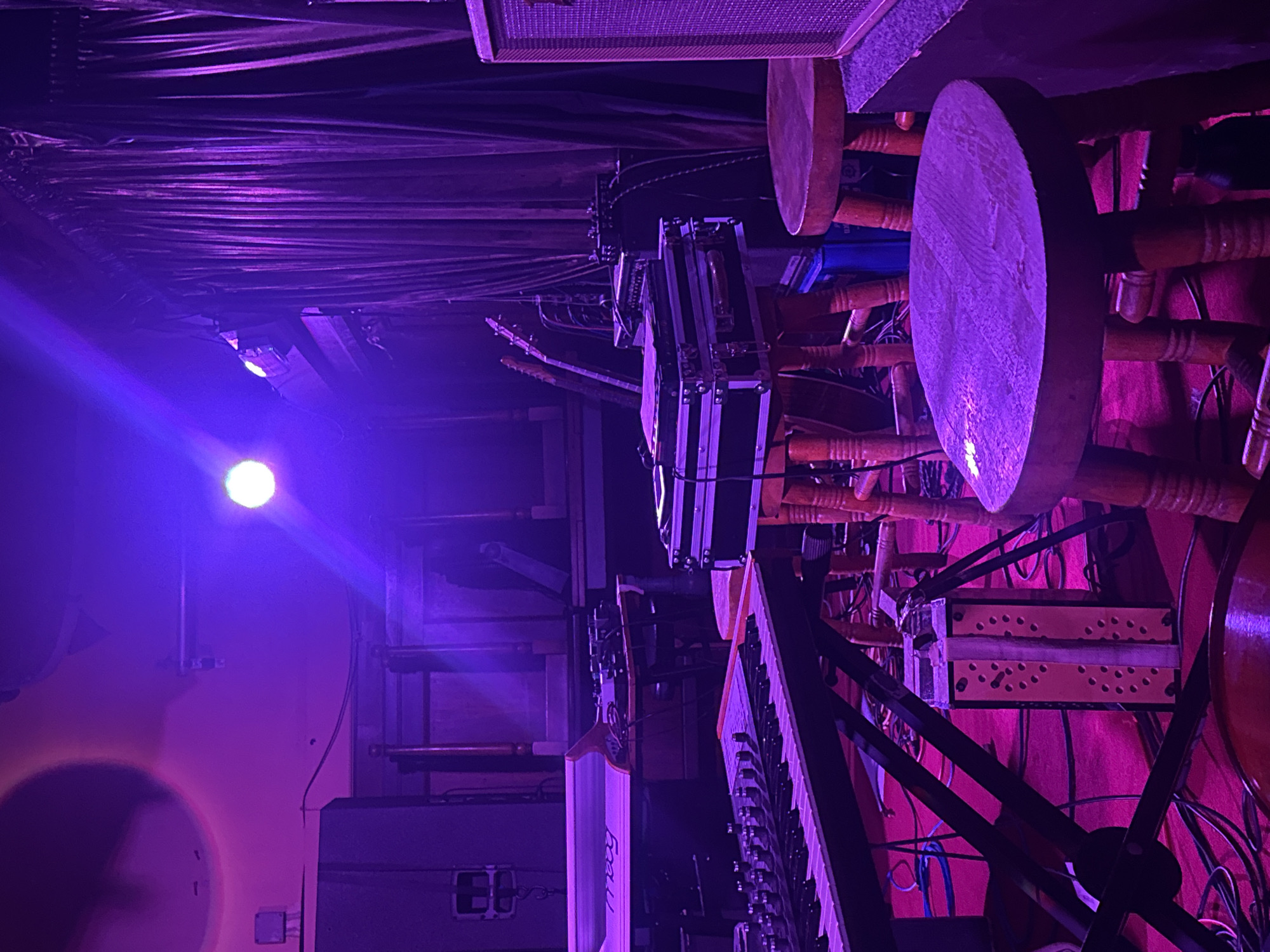 This image captures a dimly lit backstage or side-stage area of a music venue, bathed predominantly in atmospheric purple light. No people are visible in the scene.
The space is filled with musical instruments and stage equipment. On the right side of the frame, several circular, wooden drum-like instruments, possibly bodhráns, are arranged on stands, some appearing to be stacked. Below them, a black and white keyboard or synthesizer is visible, with the brand name "Moog" faintly legible on its wooden housing. Adjacent to the keyboard, multiple guitars – including at least one electric and one acoustic – rest on stands, their necks pointing upwards.
Various pieces of equipment, including what appear to be flight cases or equipment racks, are scattered around the instrument setup. Numerous cables and wires crisscross the area, connecting the different pieces of gear. Towards the top of the frame, parts of an amplifier or speaker cabinet are visible. On the far left, a bright white stage light casts a strong beam across the scene, creating a lens flare effect and illuminating the otherwise dark wall. Another amplifier can be seen on the floor in the lower-left corner. The ceiling above features textured, possibly sound-absorbing material.
The overall impression is one of anticipation, suggesting a band is either preparing for a live performance or has just finished one, set against the evocative lighting typical of a concert setting. The time of day is clearly evening or night, indicated by the artificial lighting. There are no specific visual cues within the image to suggest its location in London, United Kingdom, apart from the contextual information provided.