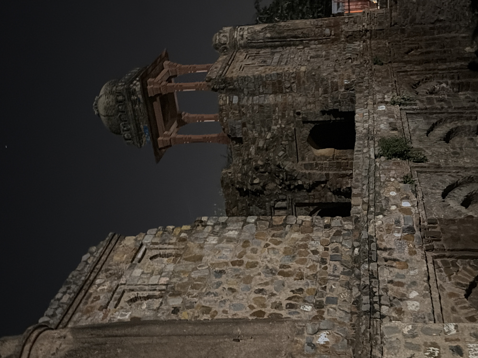 This media file depicts an old, stone structure in Delhi, India, at night. The image is taken from a low angle, looking up at the weathered stone walls and a partially ruined pavilion with a domed roof. The structure appears to be an ancient monument or fort, characterized by its rough-hewn stone blocks and architectural details like arched openings and decorative carvings.
The time of day is clearly night, with the dark sky providing a stark contrast to the illuminated portions of the building. There are no people visible in the frame, suggesting a quiet and possibly deserted setting. The weather appears clear, as there are no indications of rain or fog. The stone architecture itself is the main subject, showcasing its age and resilience through the visible wear and tear, with some areas of moss or plant growth clinging to the surfaces.
The composition emphasizes the texture and solidity of the stone, with shadows playing a significant role in defining the structure's contours. The pavilion, with its reddish-brown pillars and intricately designed dome, stands out against the darker stone of the main building. The overall mood is one of historical gravitas and a sense of timelessness. There is no visible text in the image.