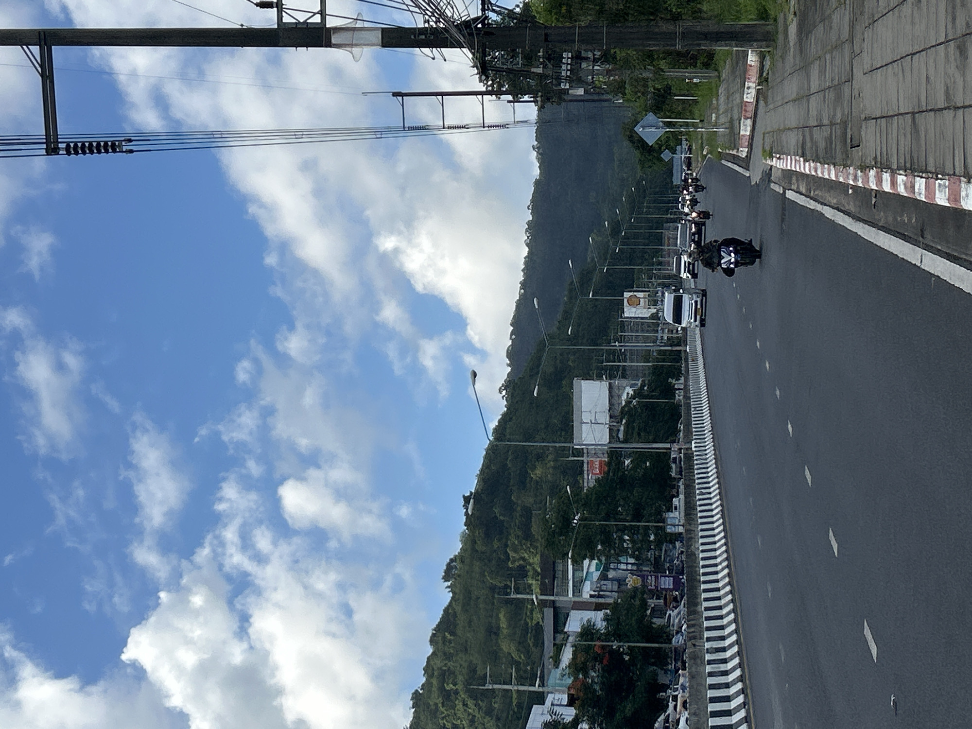 This is a medium-shot, eye-level photograph of a multi-lane road in Ratsada, Thailand, under a partly cloudy sky. The road appears to be a main thoroughfare, with concrete barriers separating the lanes and sidewalks. A row of streetlights lines the side of the road, and utility poles with numerous wires are visible on the right.
In the foreground, the dark asphalt of the road is marked with white dashed lines. On the right side of the road, a pedestrian sidewalk is visible, bordered by red and white striped curbing. A few patches of grass grow along the edge of the sidewalk.
In the middle ground, traffic is present. Several motorcycles are riding on the road, with one in the foreground appearing to be the most prominent subject, its headlights illuminated. A white van and several cars are also visible, moving in the same direction.
In the background, a densely forested hill rises behind the road, its slopes covered in lush green vegetation. Several buildings, including what appear to be commercial establishments with signage, are situated at the base of the hill, partially obscured by trees and the road infrastructure.
The sky is a bright blue, with scattered white cumulus clouds. The overall impression is of a busy urban environment integrated with a natural landscape.