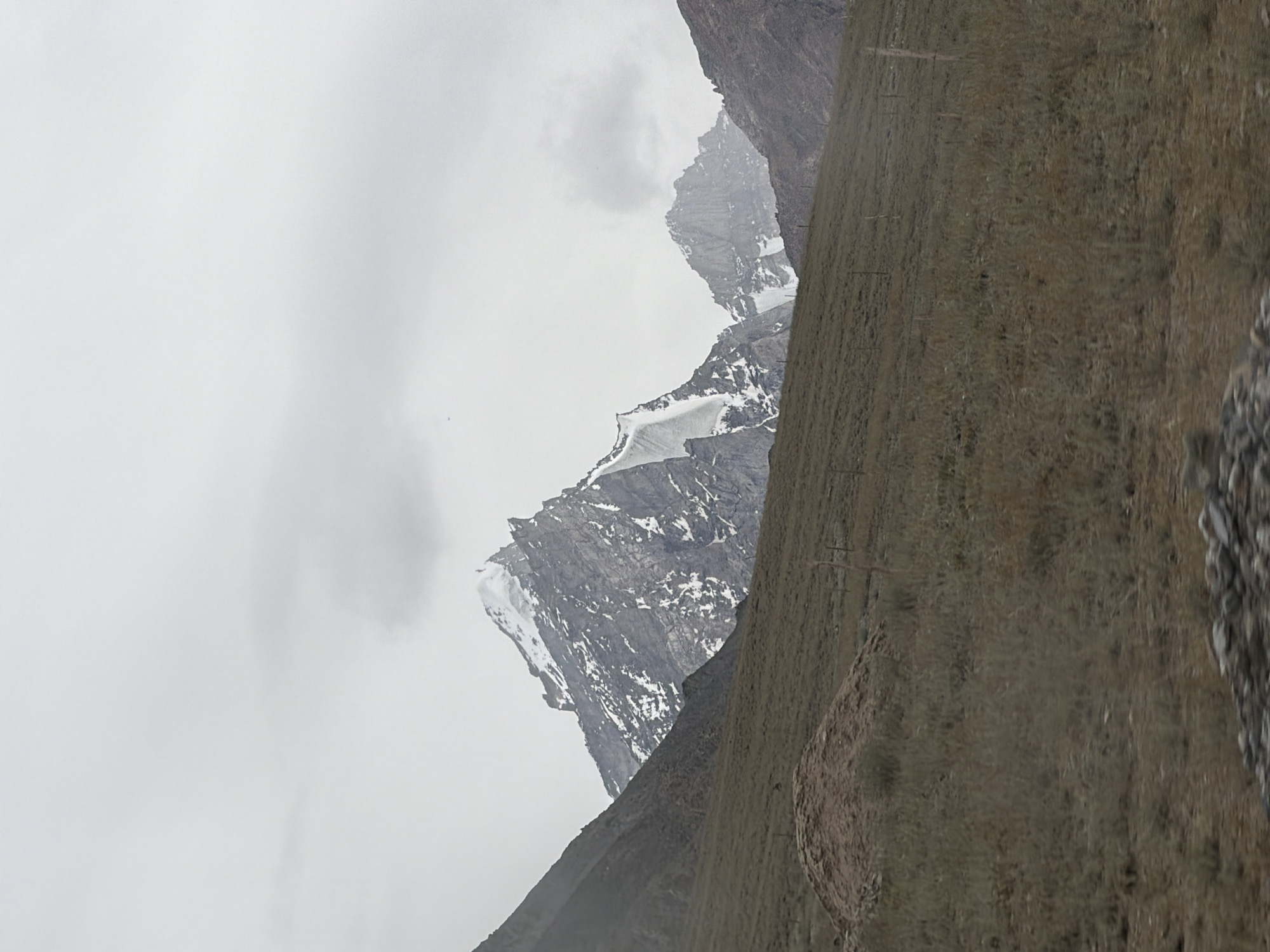A desolate mountainous landscape under a cloudy sky is depicted. The foreground is a barren, grassy slope with sparse vegetation and a small pile of soil on the left. A wire fence is visible running across the slope. In the middle ground and background, towering, jagged mountains rise sharply. The peaks are partially covered in snow, contrasting with the dark, rocky slopes. The sky is overcast and uniformly gray, with some hints of cloud formations. The scene appears to be set in a remote, high-altitude region, likely Pibiting, India, given the context. There are no people or animals present, and the overall impression is one of stark, natural beauty and isolation.