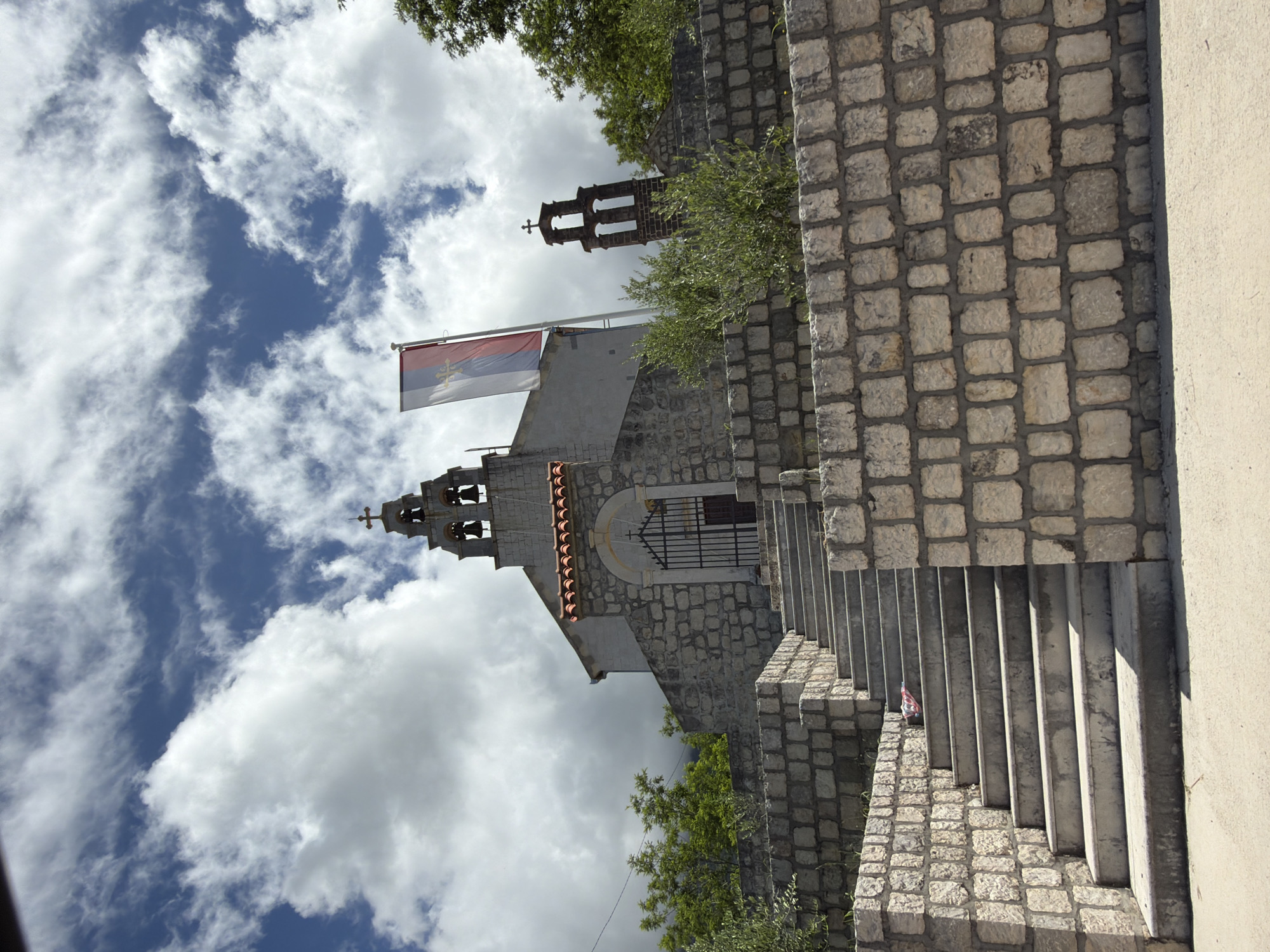 The image captures a serene outdoor scene, primarily featuring a traditional stone church or chapel nestled on a hillside under a bright, partly cloudy sky. The perspective is slightly upward, showcasing the architectural elements against the expanse of the sky.
The central subject is a stone-built church, characterized by its robust construction from irregular, light-colored stone blocks and a gabled roof covered with reddish tiles. An arched entrance, fitted with a dark, ornate wrought-iron gate, is visible on the side of the church. A prominent bell tower rises from the structure, featuring openings that house multiple bells, and is crowned with a cross. In the background, further up the hill, another, taller stone bell tower with a cross is partially visible, hinting at a larger complex or another religious edifice.
A flag flies from a pole situated near the main bell tower. It is a tricolor flag with horizontal stripes of red, blue, and white. Centered on the blue stripe is a golden emblem, which appears to be a stylized cross, possibly a Serbian cross (four C's/firesteels), indicative of historical and religious symbolism often found in the region.
To the right of the church, a robust stone wall, constructed from similarly irregular blocks, forms the base of a concrete staircase. Several concrete steps are visible, leading upwards along the right side of the image. Adjoining this stone wall on the far right is a smooth, light-colored concrete or stucco wall. Green trees and bushes are scattered throughout the scene, adding natural foliage around the stone structures.
The sky, occupying a significant portion of the image, is a vibrant blue, adorned with numerous fluffy white clouds, suggesting clear, bright daylight. No people or specific events are depicted, lending a tranquil and timeless quality to the scene. The architecture and flag strongly suggest a location in the Balkan region, specifically Bratešići, Montenegro, as provided by the context, reflecting the area's rich cultural and religious heritage.