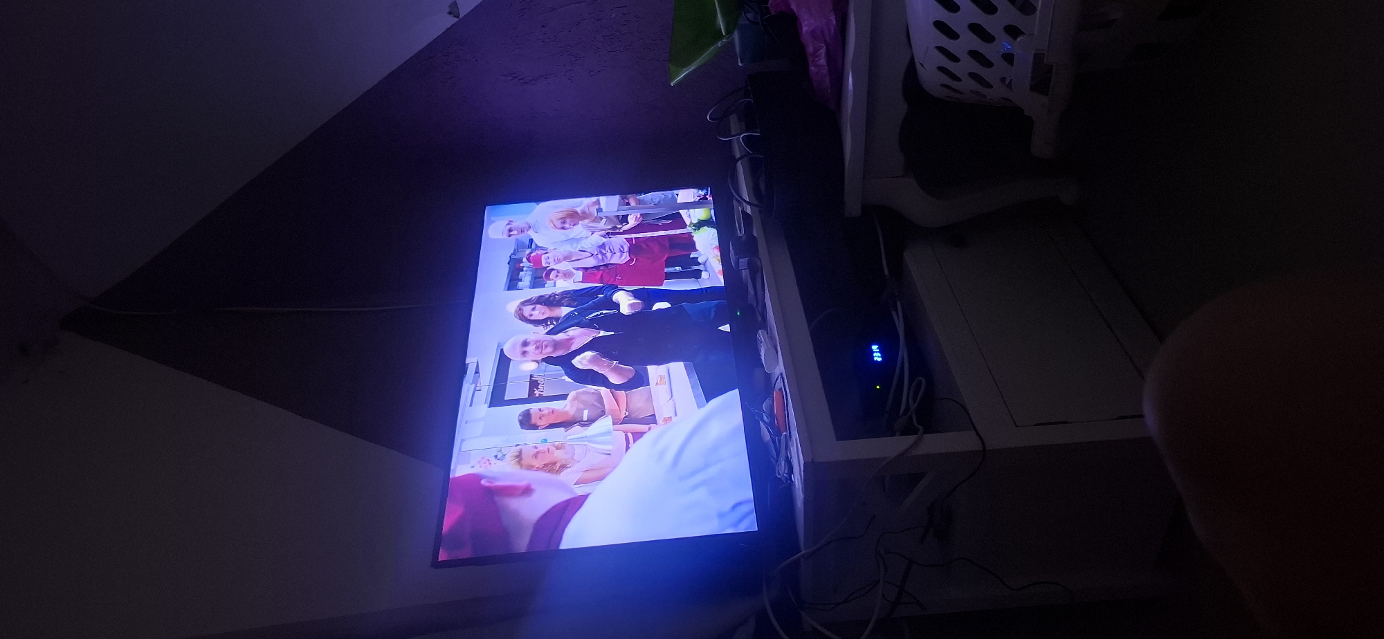 In a dimly lit room, a television screen displays a scene from a culinary show. The show features a group of people, likely contestants or participants, gathered in what appears to be a kitchen or cooking school setting. A man with a shaved head, wearing a black open-collar shirt, is the central figure on the screen, gesturing with his hands. He is surrounded by several women and another man, some wearing chef's hats or aprons. The television is placed on a white cabinet unit which also houses electronic devices, including a digital clock displaying "23:19". Cables are visible, connected to these devices. To the right of the television, another white table or cabinet holds a wicker basket and some bags. The background of the room features a dark, textured wall with a geometric pattern formed by contrasting colors, suggesting the room is decorated with a modern design. The overall ambiance is one of a domestic setting, with the television as the focal point, indicating someone is watching a program. The image was taken in Yerocham, Israel.