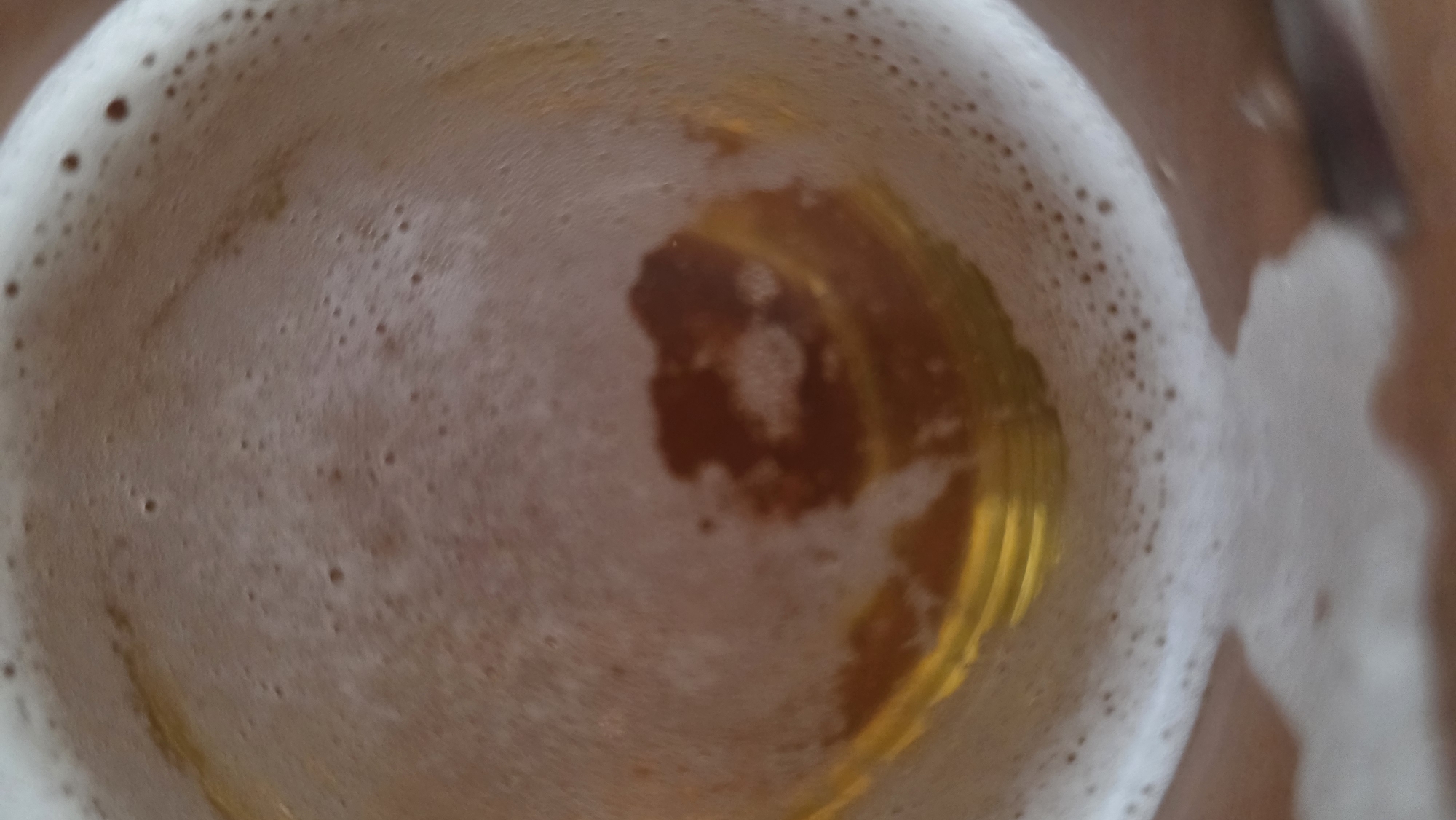 The image shows a close-up, overhead view of a glass of beer with a substantial head of foam. The beer is a light amber color. The foam is white with numerous bubbles. The inside of the glass is visible through a gap in the foam. Part of a wooden surface is visible in the upper right and bottom left.