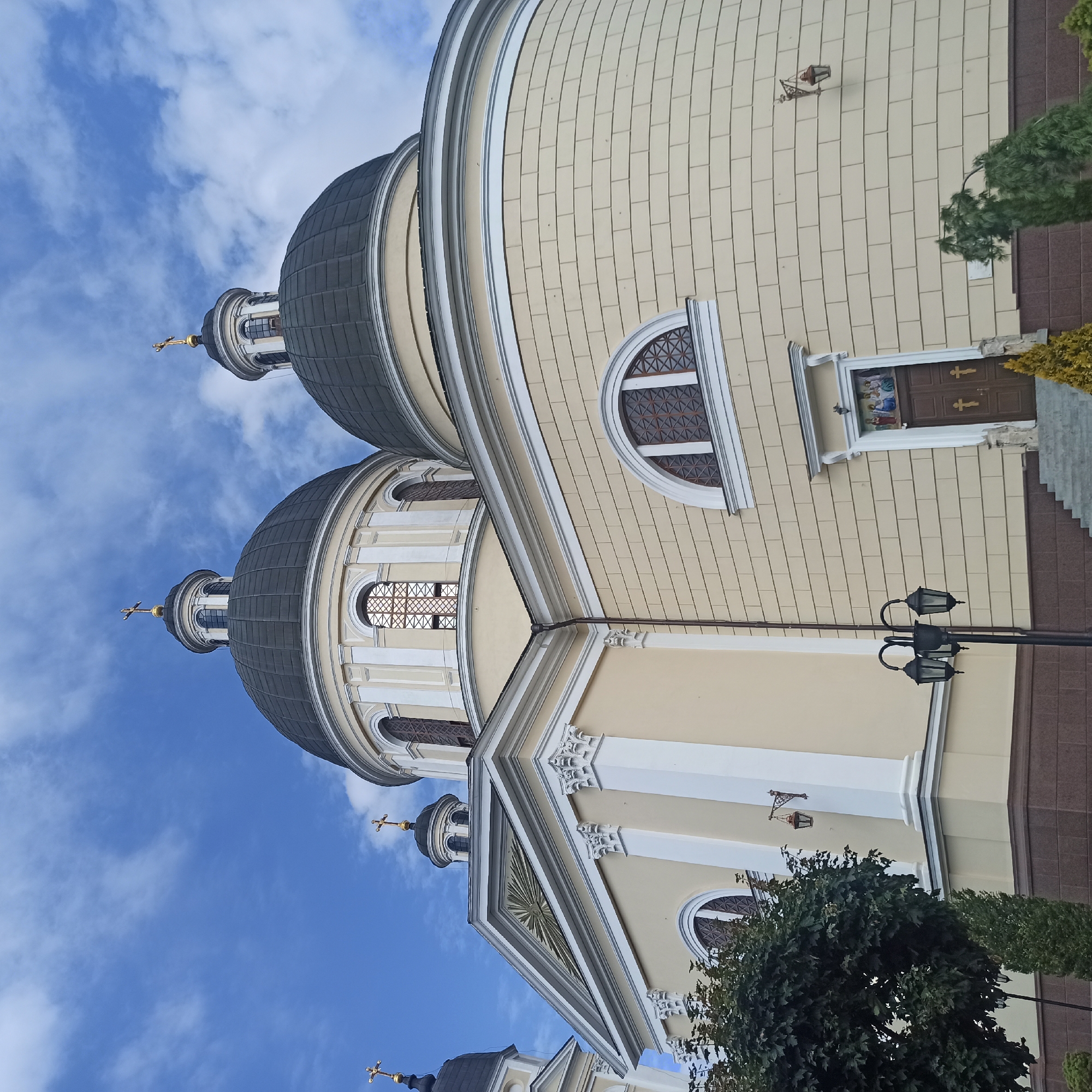 The image shows the Ascension Cathedral in Chernivtsi, Ukraine. The cathedral is a large, pale yellow building with a dark domed roof. Two large domes are visible, each topped with a cross. There are arched windows with decorative grilles. A triangular pediment with a sunburst design is visible on the left side of the building. The entrance to the cathedral is a double door with two crosses on it, above which is a religious icon. A set of stairs leads up to the entrance. There are several streetlights outside the cathedral, and some trees are visible in the foreground. The sky is blue with scattered clouds.
