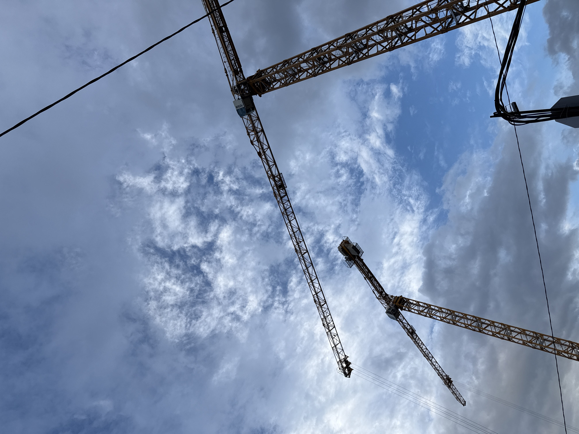 In Moscow, Russia, two yellow construction cranes are visible against a cloudy blue sky. The cranes are positioned at different angles, with one extending horizontally and the other vertically. The larger crane on the right has a cabin with "PUDIAN" written on it, suggesting it is a Potain crane. Overhead power lines are also present in the scene, crossing diagonally across the sky. The image captures a typical construction site environment with industrial machinery and infrastructure.