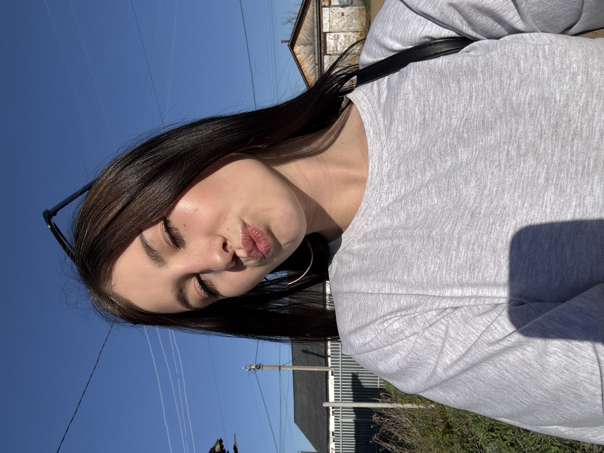 A young woman with long, dark brown hair and fair skin stands outdoors in Фоки, Russia. She is wearing a light gray long-sleeved shirt and a pair of glasses on top of her head. She has closed her eyes and is pursing her lips. A black strap goes over her shoulder. In the background, there is a blue sky with power lines, a building with a gray roof, and some greenery.