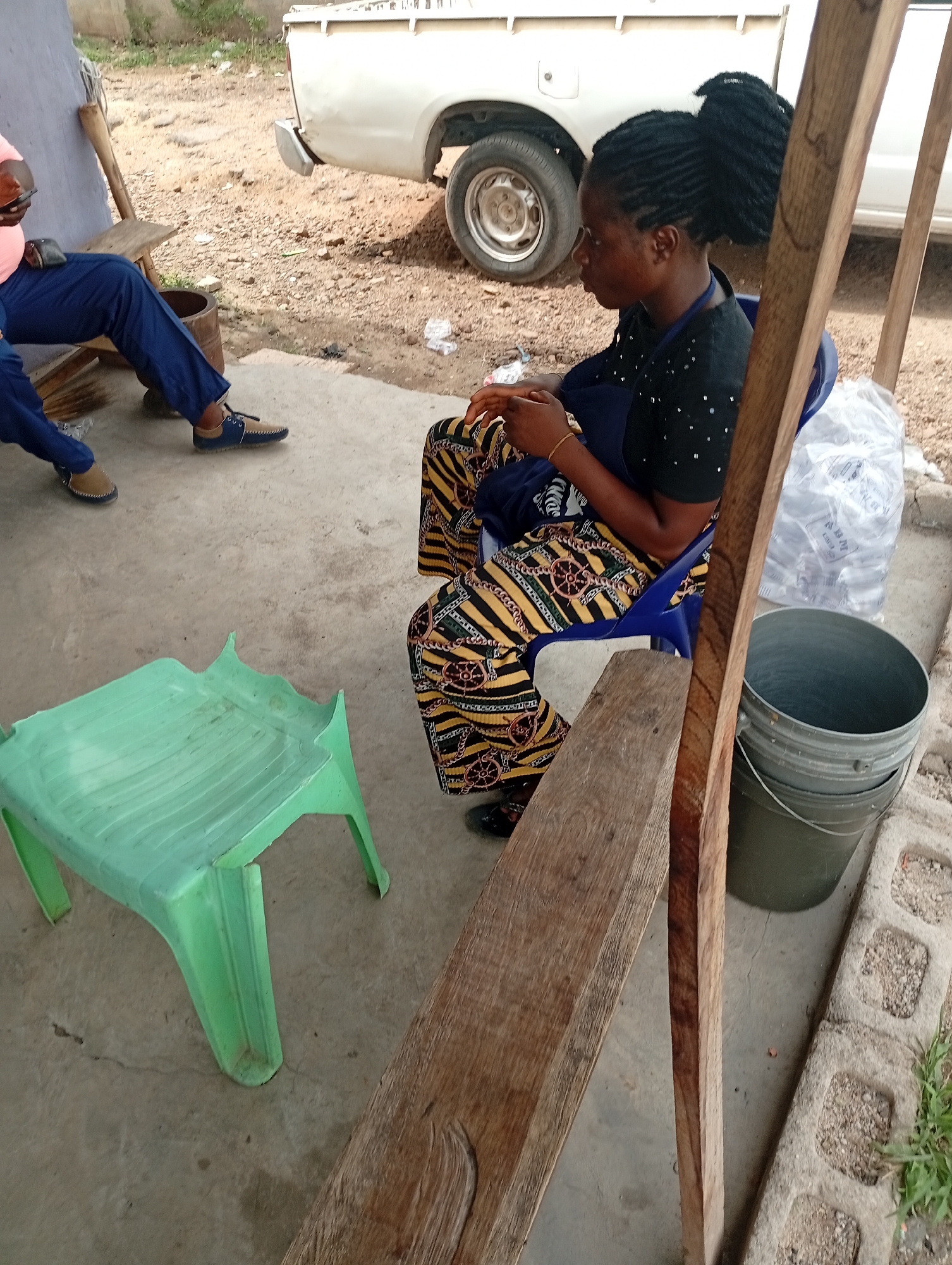 This outdoor image, captured in Adankolo, Nigeria, portrays two individuals resting in a casual setting during daylight hours. The ground in the immediate foreground is concrete, transitioning to dirt and gravel further back, where the rear of a white pickup truck is partially visible.
On the left, a man is seated on a wooden bench, wearing a pink short-sleeved top, blue trousers, and blue and tan loafers. He holds a mobile phone in his left hand.
On the right, a woman sits on a blue plastic chair, facing left in a side profile. She wears a black top with small white embellishments and vibrantly patterned trousers in yellow, black, and brown. Her dark hair is styled in braids or twists, gathered into a high bun. Her hands are gently clasped in front of her. Beside her, a grey bucket and clear plastic bags, one containing sachets of water, are visible.
A bright green plastic stool or low table is positioned on the concrete in the foreground. Several wooden planks and posts are scattered around, with one prominent wooden beam running vertically on the right, partially framing the scene. The overall atmosphere suggests a relaxed, informal gathering or waiting area. No discernible text is visible on any objects.