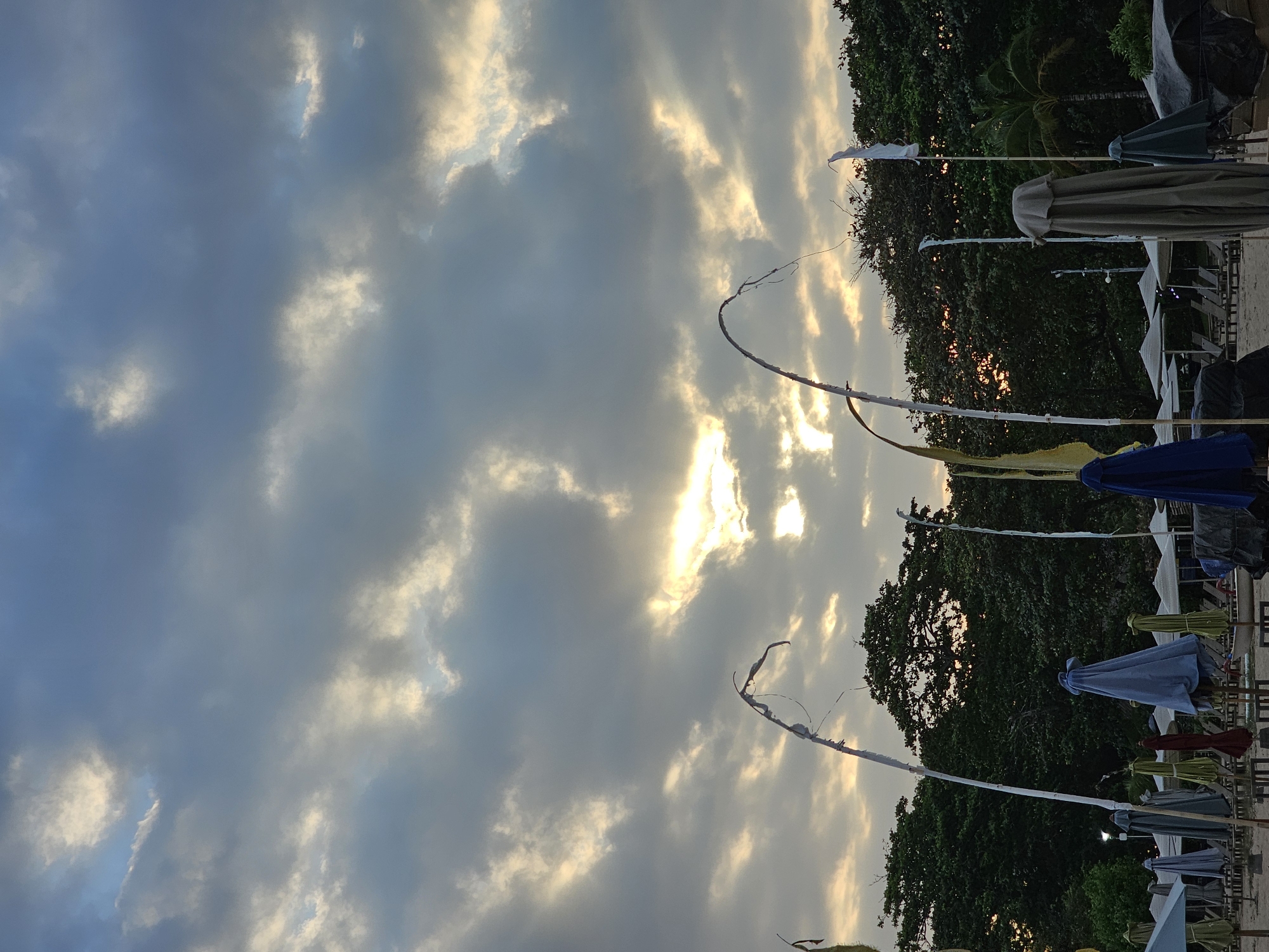 The image captures a serene beach scene in Denpasar, Indonesia, under a dramatic, cloudy sky. The sun is partially visible, breaking through the clouds and casting a golden glow. In the foreground, several closed beach umbrellas stand in rows on the sandy shore. Tall, thin poles with white and yellow streamers are interspersed among the umbrellas, some of which are covered with fabric. Behind the umbrellas, a dense line of lush green trees forms the backdrop, with the setting or rising sun peeking through their branches. The overall impression is one of tranquility and natural beauty, with the man-made elements of the beach furniture blending into the environment.
