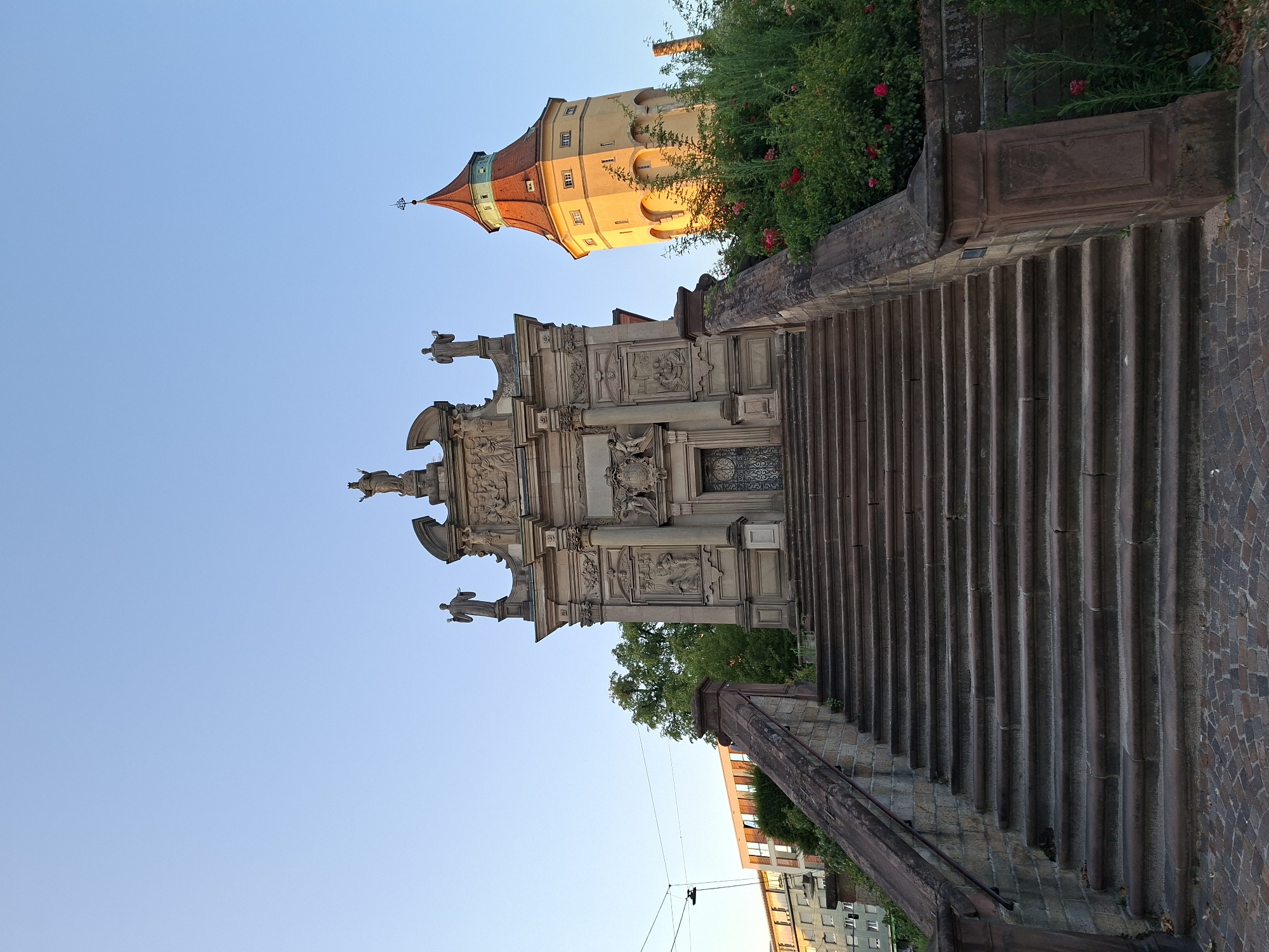 The image captures the "Fideliskapelle" and the associated water tower in Rastatt, Germany, bathed in the warm light of what appears to be sunset or sunrise. A grand stone staircase leads up to the chapel, which is an ornate Baroque structure adorned with sculptures and architectural details. To the right of the chapel, a distinctive yellow water tower with a red-tiled, pointed roof stands prominently. Lush green bushes and red roses surround the base of the water tower and climb along the side of the staircase, adding vibrant color to the scene. In the background, a modern apartment building with a light-colored facade and visible windows is visible, along with utility wires and a street lamp hanging from them, indicating an urban setting. The sky is a clear, pale blue, suggesting fair weather conditions. No people are visible in the frame.
