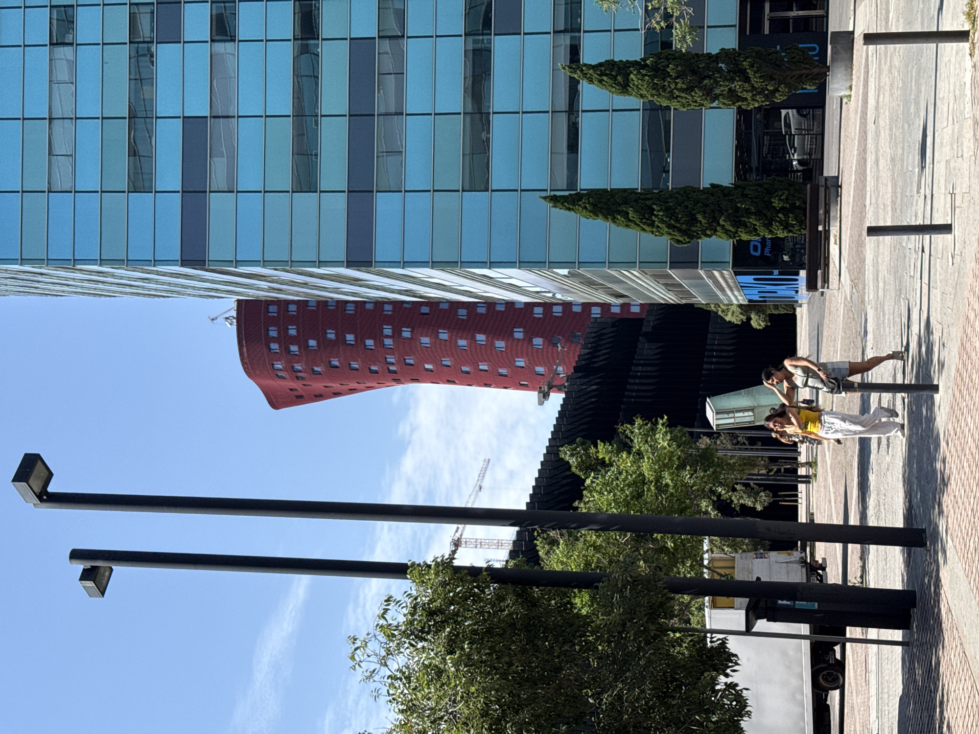 In L'Hospitalet de Llobregat, Spain, two women are walking on a paved sidewalk next to a street. The woman on the left wears a yellow tank top and white pants, while the woman on the right wears a green tank top and denim shorts. They are both looking towards the left. In the background, several modern buildings are visible, including a tall, blue glass skyscraper and a distinctive curved red building. Trees line the sidewalk, and streetlights are present. A white truck is parked on the left side of the image. The sky is clear and blue with some clouds.