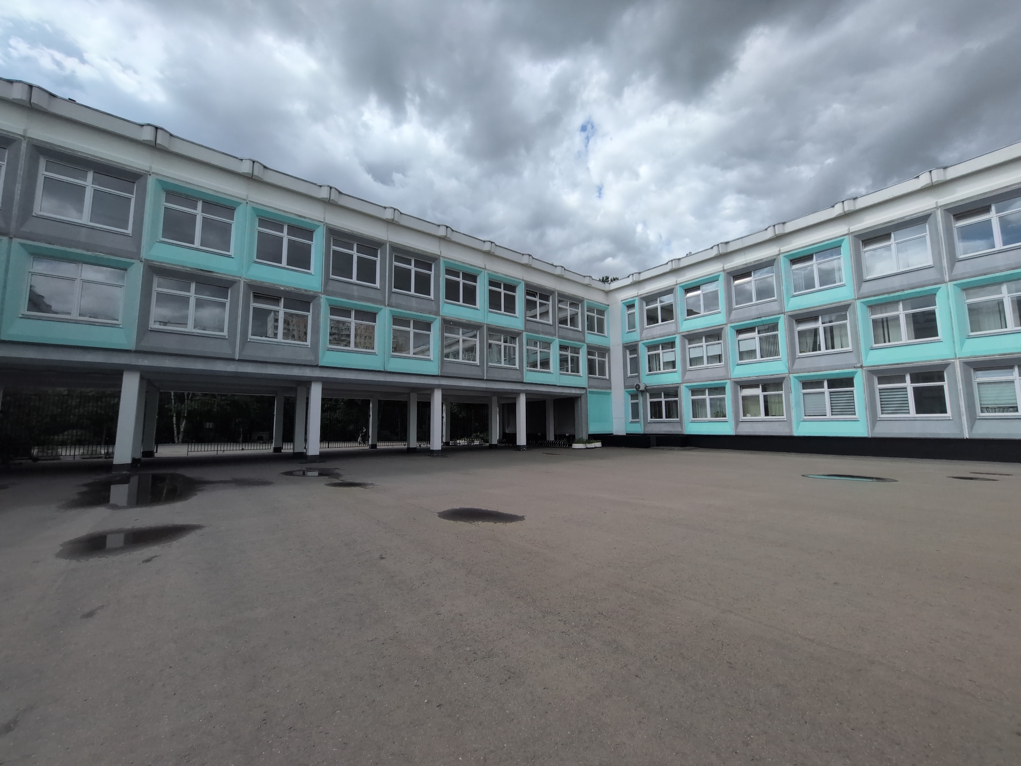 The image shows the courtyard of a building, possibly a school, in Moscow, Russia. The building is a three-story structure with a repetitive pattern of windows framed in grey and aqua. There are support pillars on the left side creating a covered walkway. The courtyard surface is paved with asphalt, and there are puddles on the ground. The sky is overcast with heavy, grey clouds. No people are visible in the image.