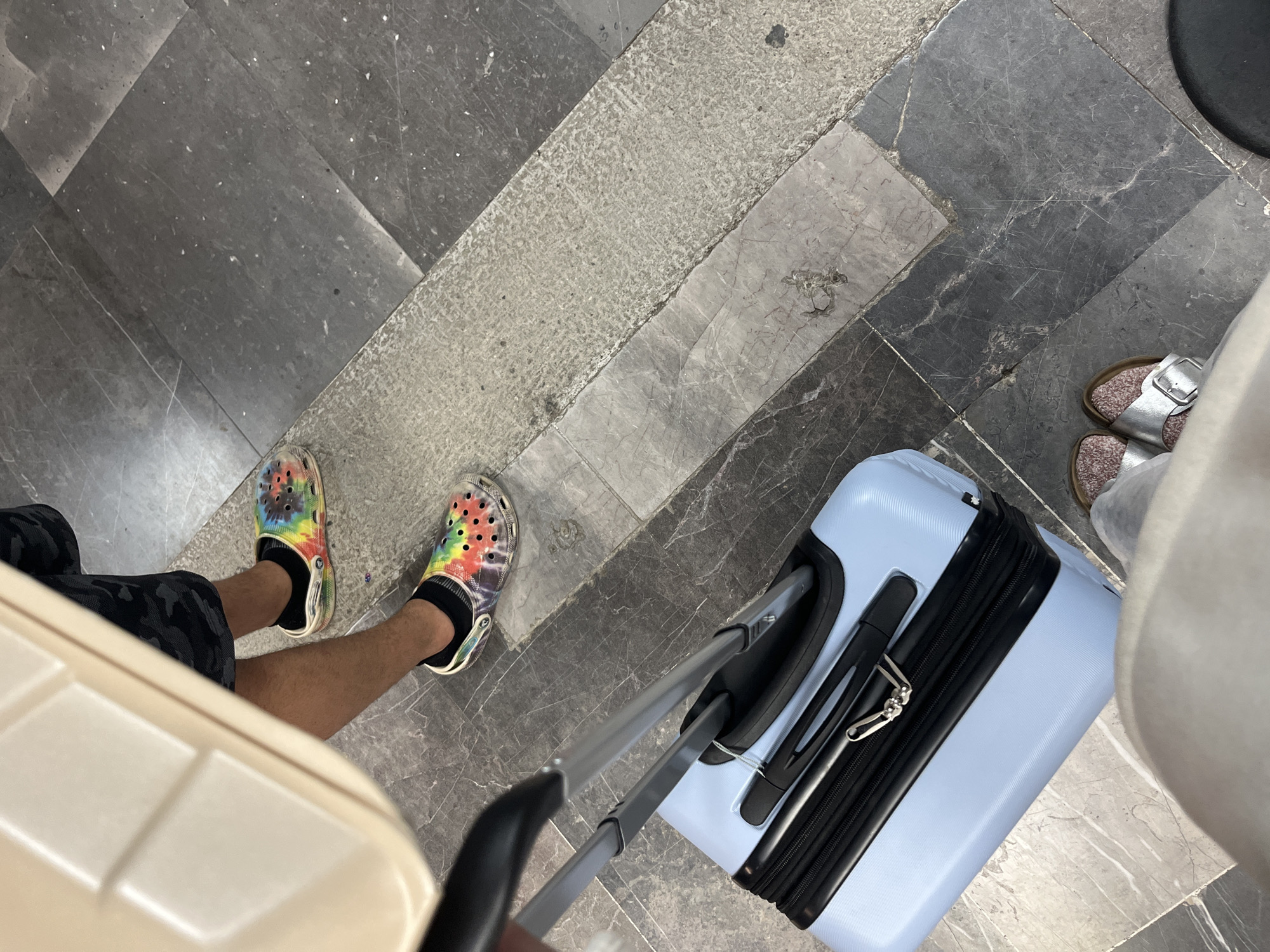The image depicts a first-person perspective view of two individuals at an airport or transport hub. One individual is wearing tie-dye Crocs with black socks, camouflage shorts, and is pulling a beige suitcase. The other person is wearing silver sandals, light pants and appears to be pulling a light blue suitcase. The floor is composed of dark and light gray marble tiling with a concrete pathway. The specific location in Puerto Escondido, Mexico, is not discernible from the provided image.