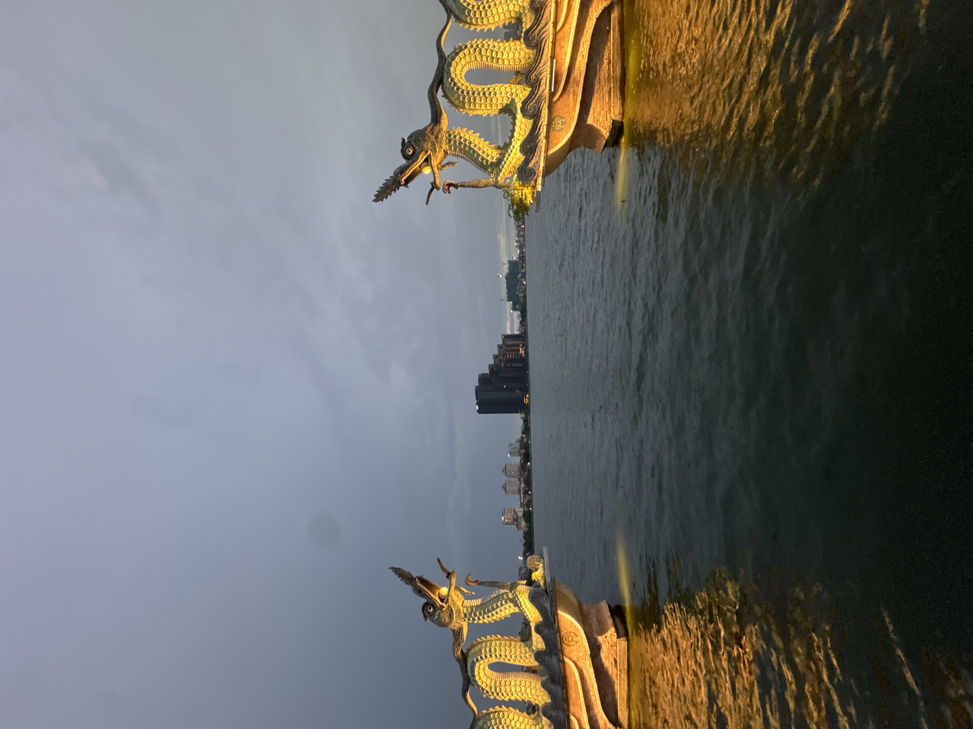 An outdoor, eye-level, wide shot captures the twilight scene at West Lake in Hanoi, Vietnam. The sky is overcast with dark gray clouds. In the foreground, two illuminated dragon sculptures, one on the left and one on the right, arch over the water. These sculptures are yellow and gold, with detailed scales and open mouths, appearing to be part of ornate boat prows. The water below is dark green and choppy, reflecting the warm yellow light from the dragon sculptures. In the middle ground, a body of water stretches towards a cityscape visible in the distance. The cityscape features several tall, dark buildings and other lower structures, with construction cranes also present. The overall atmosphere is dramatic and serene, with the contrasting light and dark elements emphasizing the architectural features against the natural environment.