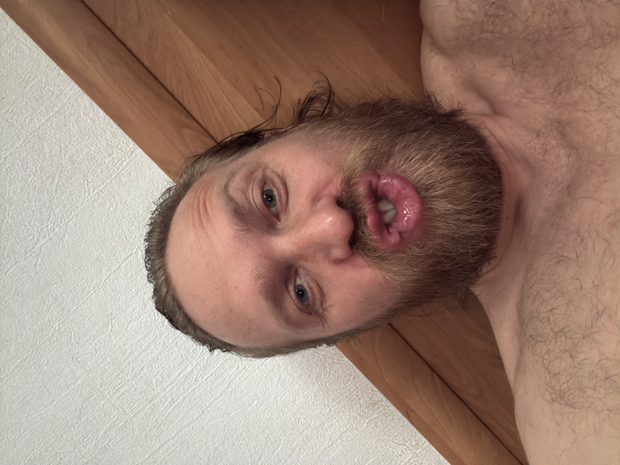 A man with a beard is photographed close-up, lying down. His face is turned toward the camera, and he is making a face with his mouth open. Part of his bare chest and shoulder are visible. The background consists of a white textured wall and a wooden surface. The location of the scene is not discernible from the image.