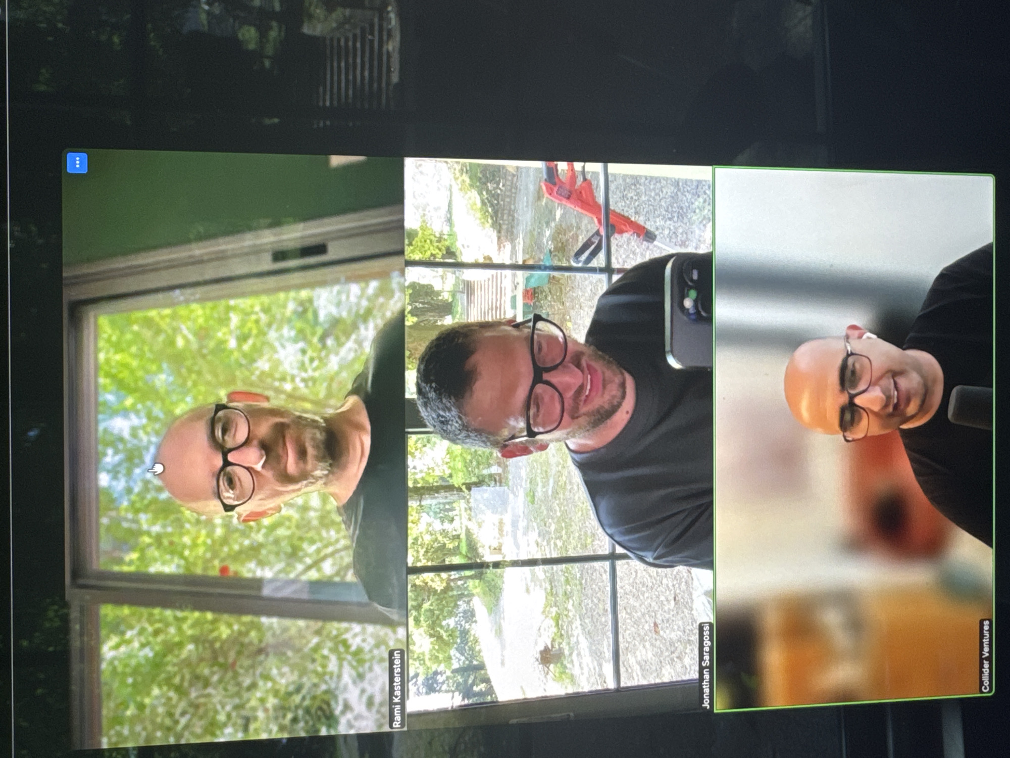 The image shows a screen displaying a video conference with three participants. Each participant occupies a separate panel. The participant on the left is named Rami Kasterstein and is positioned in front of a window with a view of greenery. The middle participant, Jonathan Saragossi, is holding a phone up to his face, revealing a view of an outdoor patio area. The participant on the right represents Collider Ventures. All three participants are bald men wearing glasses and black shirts. The location is unlikely to be Erba, Italy given the attire and participants.