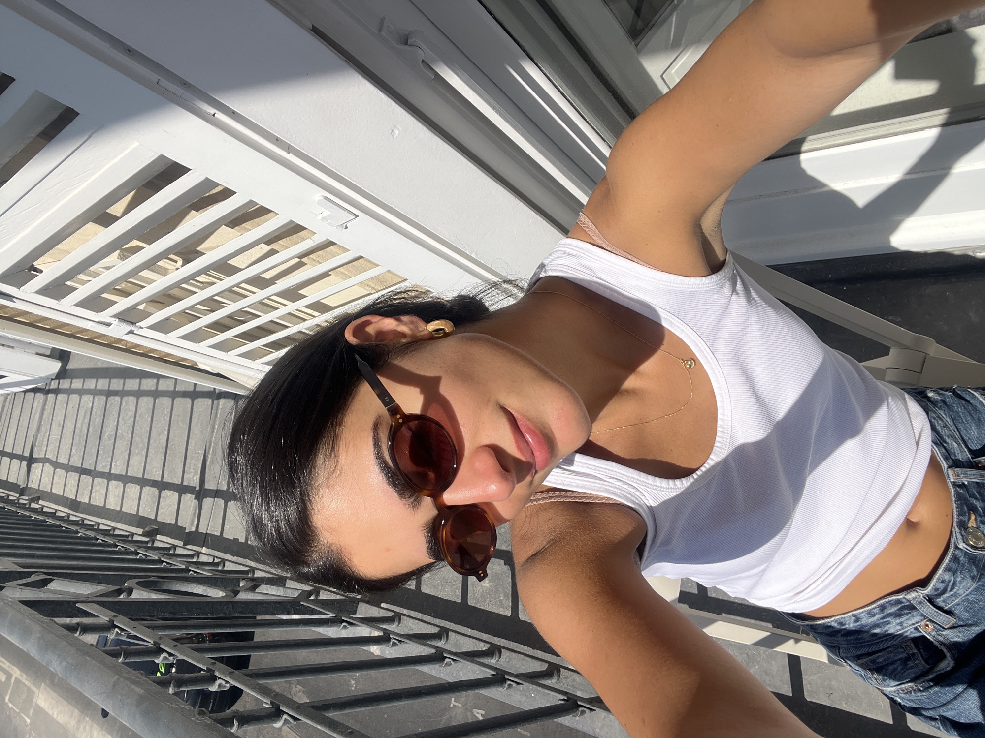 A woman takes a selfie outdoors with buildings in the background. She has dark hair, sunglasses, a white tank top and jeans. She is standing on a balcony with a metal railing. White-painted latticework is visible above. The photo appears to have been taken in Paris, France.