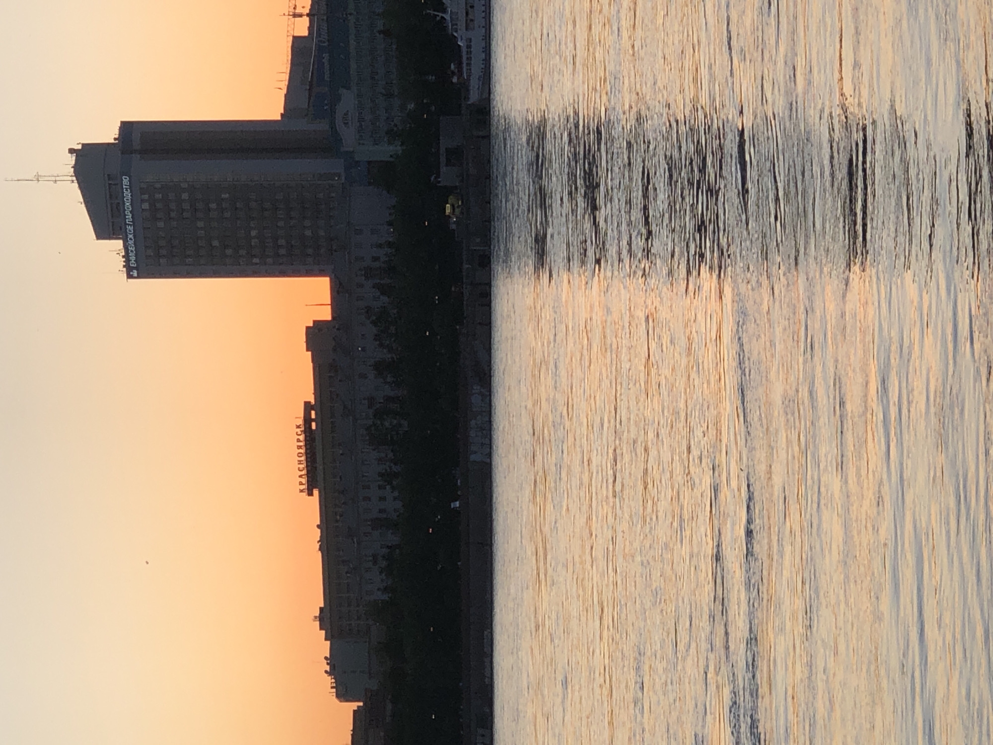 The image captures a serene cityscape view along a river during the golden hour, likely sunrise or sunset, in Krasnoyarsk, Russia. The sky, occupying the left portion of the frame, transitions from a soft orange and yellow at the horizon to a paler hue higher up, indicating clear weather.
Silhouetted against the vibrant sky, a line of buildings forms the city's profile. The most prominent structure is a tall, dark skyscraper, standing out distinctly. On its upper facade, Russian text is clearly visible, reading "ЕНИСЕЙСКОЕ ПАРОХОДСТВО" (Yenisei Shipping Company). Further along the riverbank, on a lower building, another sign spells out "КРАСНОЯРСК" (Krasnoyarsk) in large letters. The buildings appear mostly in shadow, emphasizing the dramatic sky and water reflections.
The right side of the image is dominated by the river, which beautifully reflects the warm, golden light of the sky. The water's surface is characterized by numerous vertical streaks of light and shadow, creating a textured, almost painterly effect from the gentle ripples. A darker, more pronounced horizontal band of reflections cuts across the mid-section of the river, contrasting with the lighter, shimmering areas.
The scene conveys a peaceful atmosphere. No people or specific activities are visible, allowing the natural light and urban architecture to be the primary subjects. The visible text on the buildings explicitly confirms the location as Krasnoyarsk, Russia, alongside the Yenisei River.