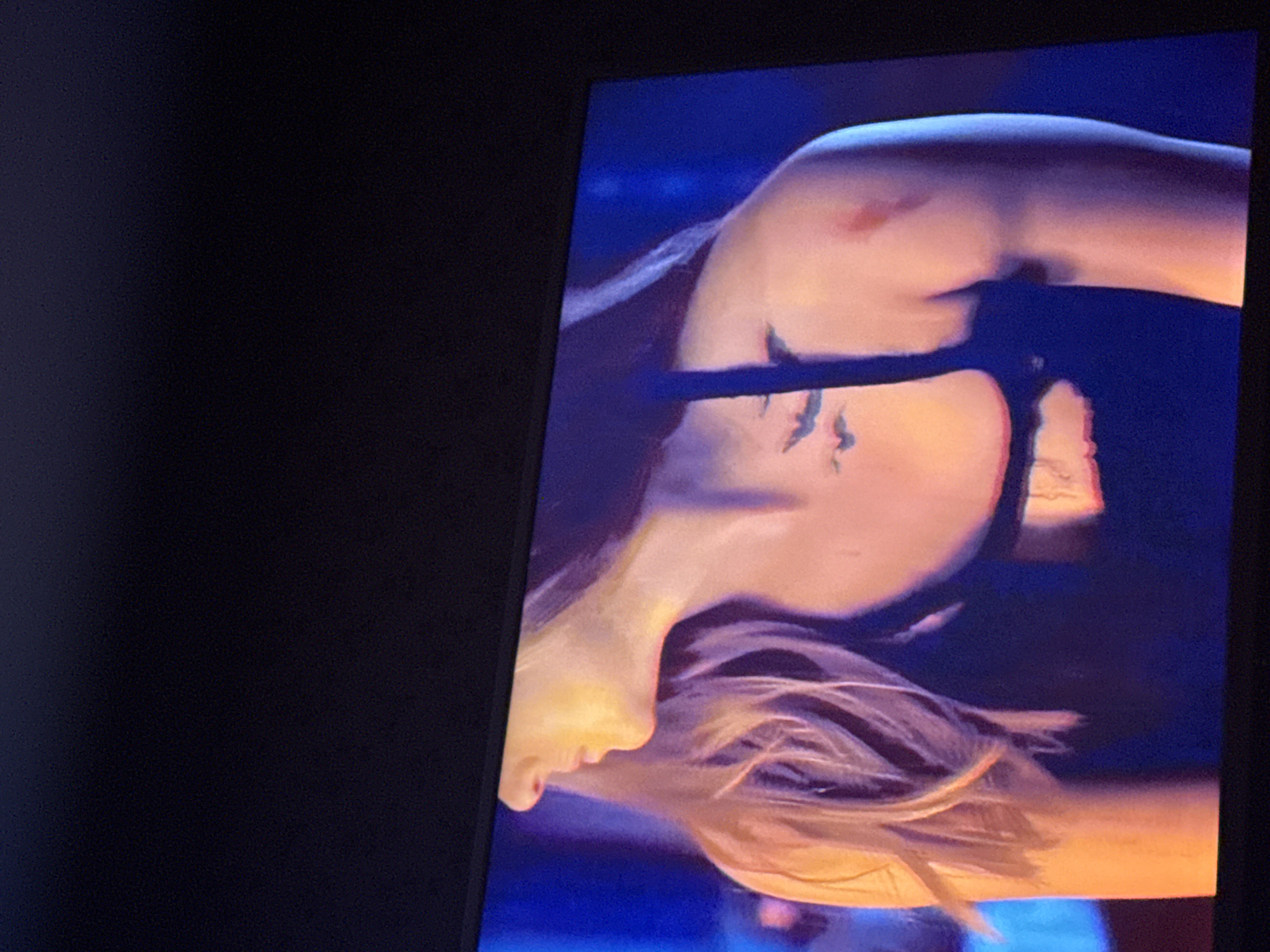 The image is a close-up photograph of a person's upper body, likely viewed on a screen. The person has fair skin and light-colored hair. The image is rotated, with the person's face in the lower left corner and their shoulder in the upper right. There is a black strap visible across their chest and what appears to be a tattoo of birds above the strap. The background is a blurry mix of blue and orange hues. The photograph itself seems to be taken of a monitor or screen due to the surrounding black frame. Location details for Mytishchi, Russia are not provided in the image.