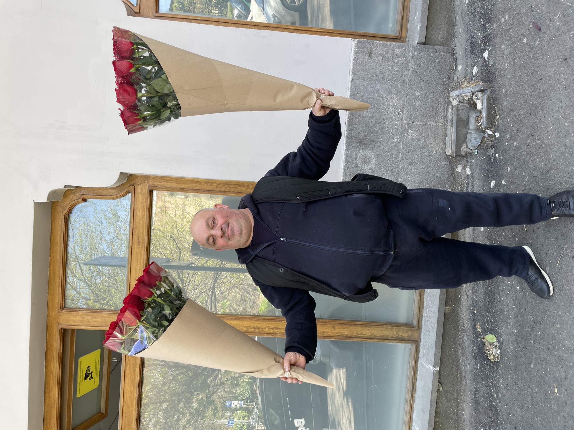 A bald, middle-aged man stands outdoors, holding two large bouquets of red roses. He is dressed in a dark blue or black zippered jacket, dark pants, and black sneakers, and wears a slight smile. Each bouquet is wrapped in light brown kraft paper and clear plastic, showcasing the vibrant red blossoms. He holds one bouquet in each outstretched hand.
The man is positioned on a grey, weathered concrete or asphalt sidewalk. Behind him is a building with large, ornate wooden-framed windows that reflect green trees, a clear blue sky, and what appears to be a car or other urban elements. One window pane contains a yellow square sign with a black security camera icon and some small, illegible black text. Near the man's right foot (viewer's left), an old, damaged metal grate or drain is embedded in the pavement, surrounded by dirt and debris. Faint, partial white text, possibly "Böl", is visible on the ground.
The scene takes place during the daytime under bright, possibly sunny, weather conditions, consistent with an urban setting like Tbilisi, Georgia. The man appears to be posing for a photograph, presenting the flowers.