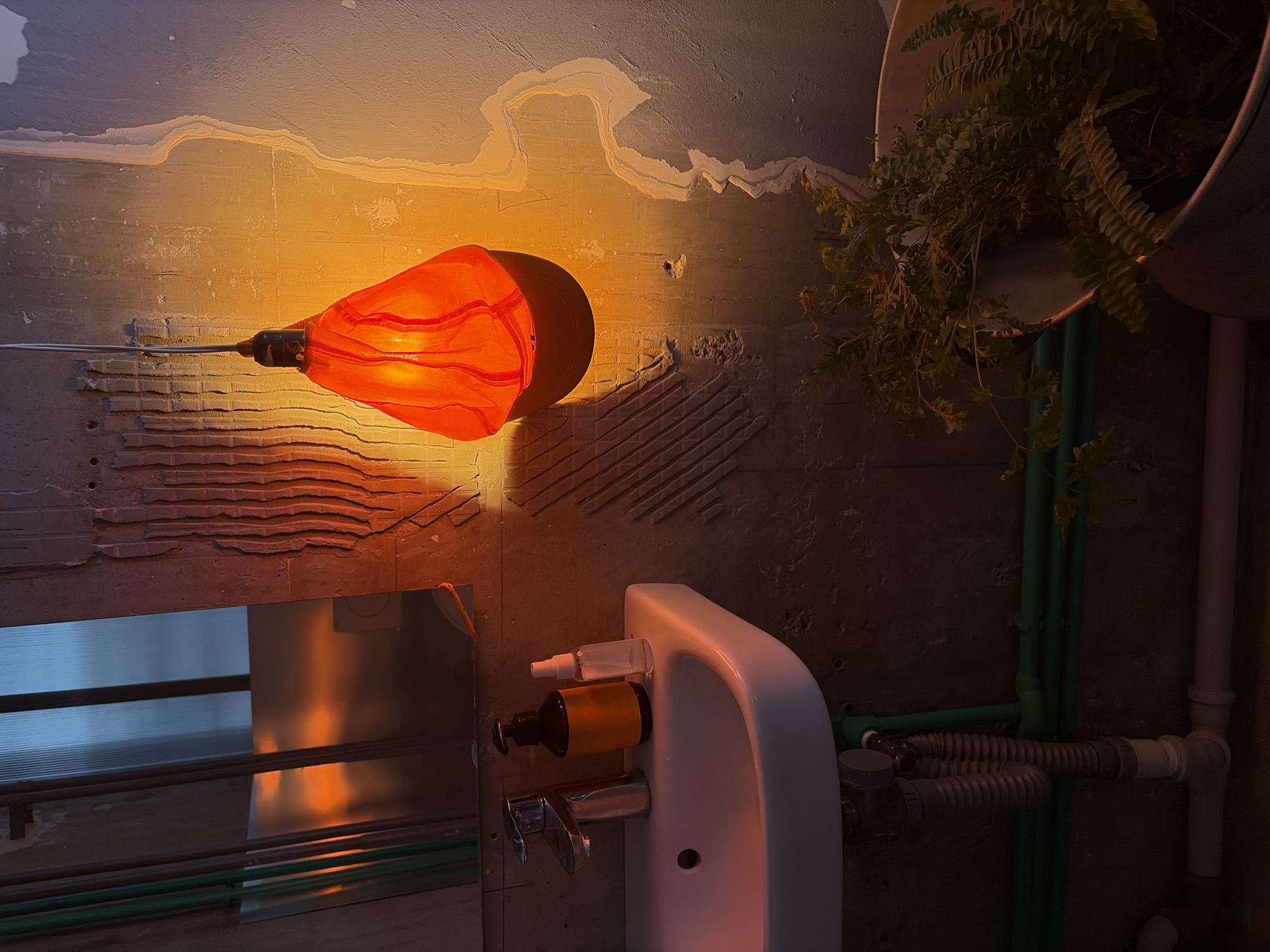 The image portrays a dimly lit interior space, likely a bathroom or utility area, characterized by an industrial aesthetic with exposed concrete walls. An orange glass pendant lamp, featuring a striped pattern, is suspended from the left, casting a strong, warm amber glow across the scene.
The concrete walls display varied textures; some areas show peeling paint or plaster, revealing underlying layers, while others have rough or textured patterns, possibly from removed finishes. In the upper right, a green fern plant in a light-colored pot hangs, partially illuminated by the lamp and partially in shadow.
Below the lamp, a horizontal mirror or polished metal sheet reflects the intense orange light. To the right, a white, wall-mounted sink with a silver faucet is visible. Attached to the wall above the sink are two dispenser bottles: a clear spray bottle and an amber pump bottle.
Below and to the right of the sink, plumbing pipes in green, white, and grey corrugated materials run along the concrete wall. The overall impression is one of a functional, possibly artistic or under-construction environment, strongly illuminated by the artificial light, suggesting evening or night. No text is visible in the image. The location is contextualized as Belgrade, Serbia.