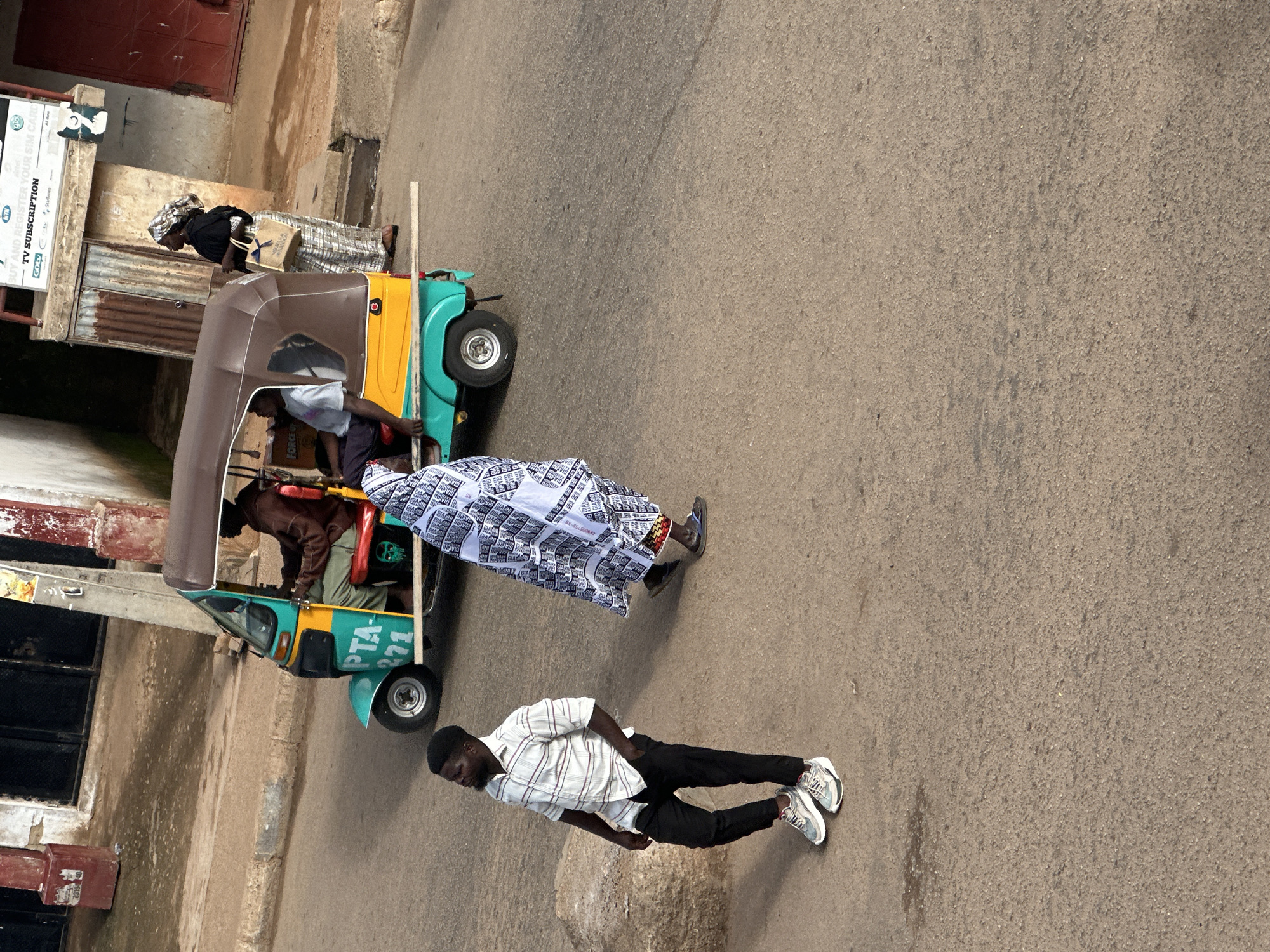 In Bukuru, Nigeria, a three-wheeled motorized vehicle, commonly known as a "keke," is parked on a paved street. A person is seated in the driver's position of the keke, wearing a brown hoodie. Another person is visible inside the keke, wearing a light-colored t-shirt. A woman stands to the right of the keke, dressed in a black top, a headscarf, and a patterned skirt, carrying a light-colored bag. In the foreground, a man walks away from the camera, wearing a checkered short-sleeved shirt, black trousers, and athletic shoes. To the left of the man, a large rock is situated on the side of the road. Another person, draped in a white and black patterned cloth, walks away from the camera towards the keke. The background features buildings with corrugated metal roofing and windows, and a street sign displaying "TV SUBSCRIPTION." The ground surface is asphalt, with some dirt and debris.