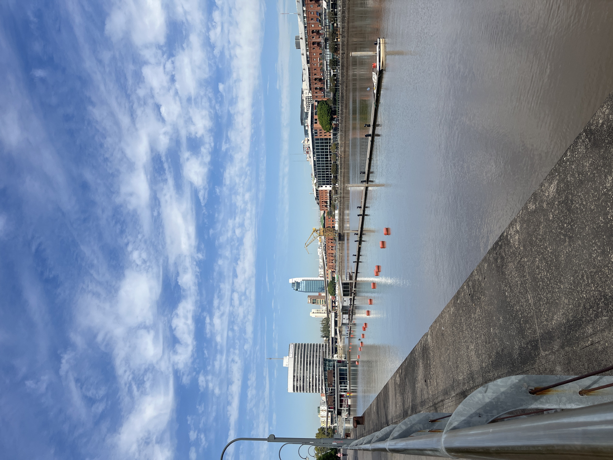 The image shows a view from a bridge or elevated walkway in Buenos Aires, Argentina, overlooking the harbor area. The sky is blue with wispy clouds. The water is calm with reflections of the buildings and sky. In the distance, there are modern buildings and some older brick structures. A yellow construction crane is visible. There are floating orange buoys in the water. A concrete barrier is in the foreground, with metal railings visible.