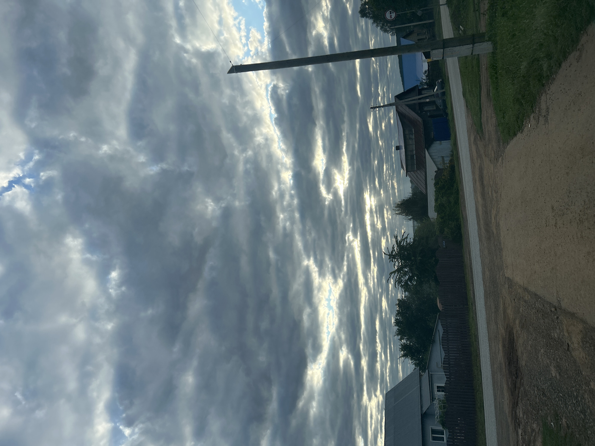 The image depicts a rural landscape under a dramatic, overcast sky. The clouds are heavy and dark, with streaks of light breaking through, suggesting either sunrise or sunset. In the foreground, a dirt road leads towards a paved road. To the left, a light blue house with a grey roof is visible, bordered by a dark wooden fence. Further down the paved road, several houses with traditional wooden architecture are scattered, some with dark roofs and others with blue roofs. Dense green trees and bushes are present throughout the scene, indicating a natural environment. A tall wooden utility pole stands prominently on the right side of the image, with wires running across the frame. A traffic sign with a circular red border and a diagonal bar is also visible on the right. The overall impression is one of a quiet, possibly remote, village setting in Фоки, Russia, characterized by its natural beauty and simple architecture. There are no people visible in the image.