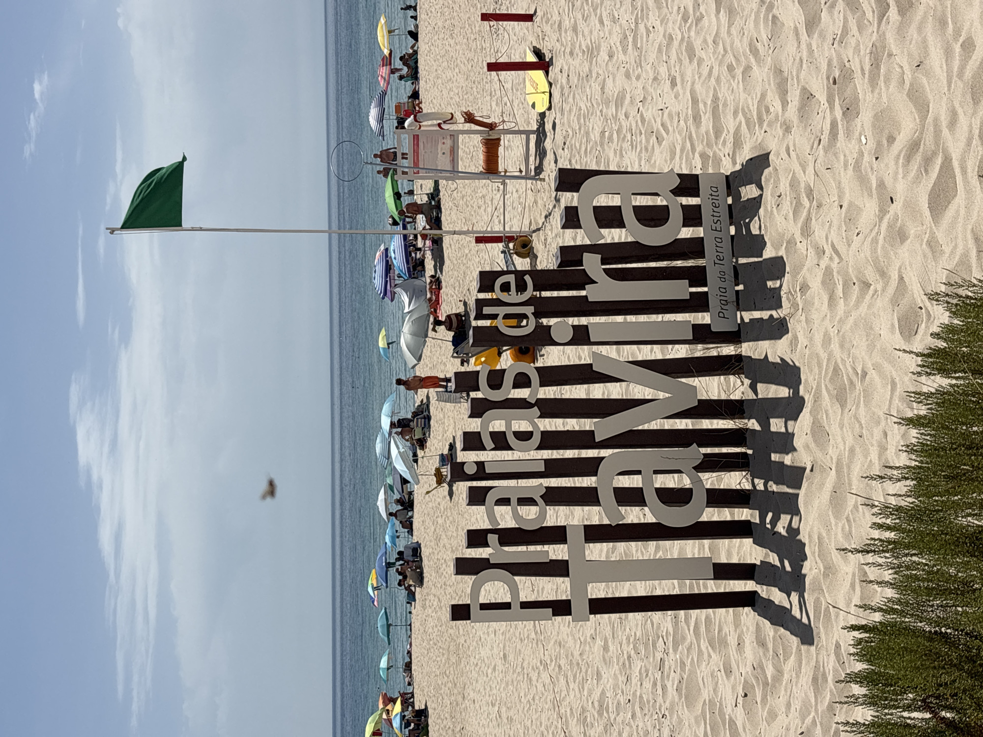 A wide shot captures a beach in Tavira, Portugal, on a bright, sunny day. In the foreground, a large wooden sign with white lettering spells out "Praias de Tavira," with a smaller sign below reading "Praia da Terra Estreita." The beach is covered in fine, light-colored sand.
In the mid-ground, several beach umbrellas in various colors are spread out, with people relaxing underneath them or walking on the sand. The ocean, a deep blue, stretches to the horizon. A green flag flies from a tall flagpole, indicating safe swimming conditions. A lifeguard stand with a life preserver and rope is visible near the center of the frame, suggesting a monitored beach. A yellow surfboard lies on the sand near the lifeguard stand.
In the background, the distant shoreline is visible, and the sky is a clear blue with a few wispy white clouds. A bird is captured in mid-flight in the upper left portion of the image. Green shrubs line the bottom edge of the frame, partially obscuring the sand.