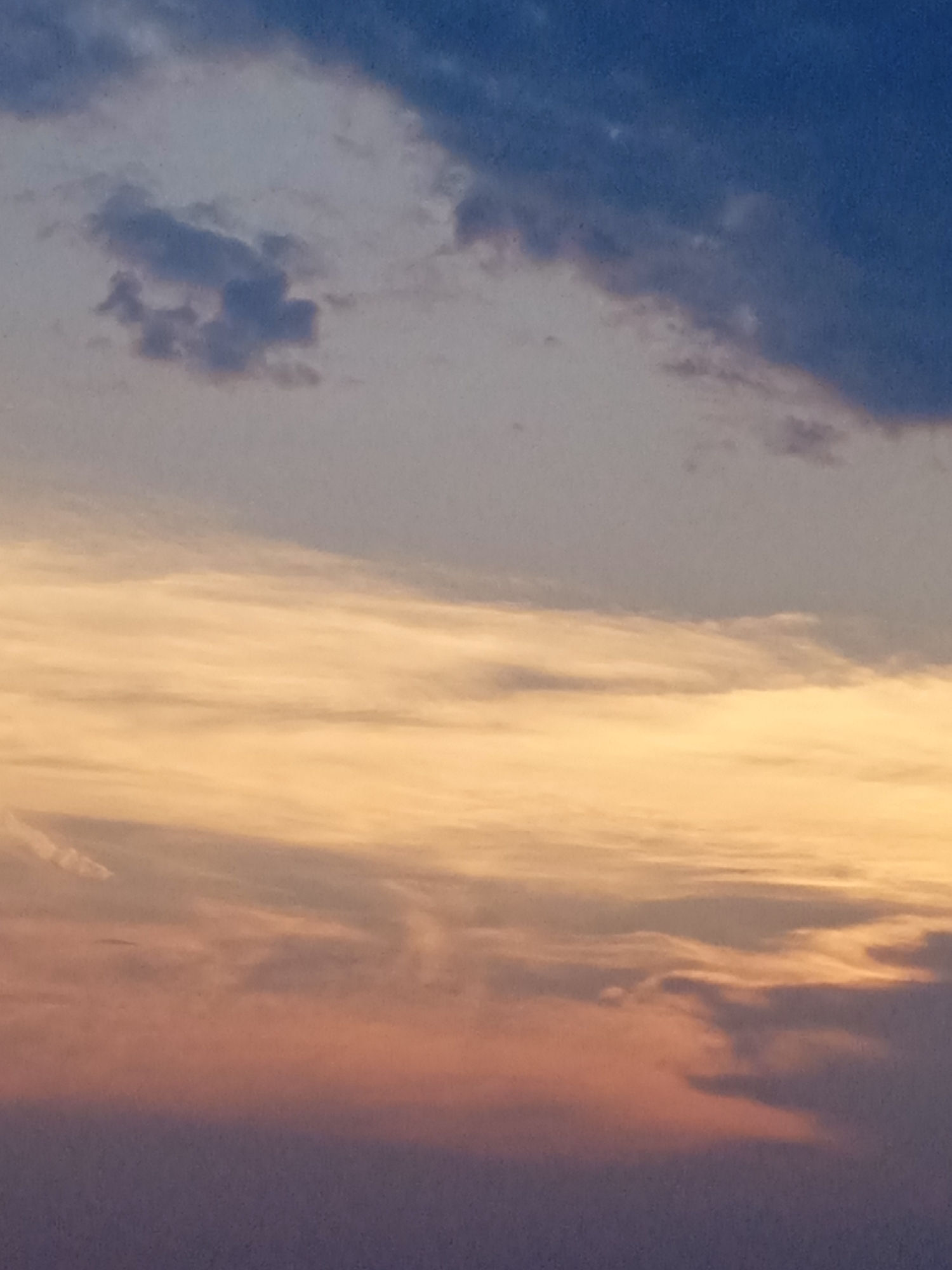 The image shows a sunset sky with layered clouds. The top of the image features dark blue clouds, transitioning to lighter gray and then to a broad band of warm orange and yellow clouds. Towards the bottom, the clouds are a muted pink and purple, fading into a dark, blurry foreground that could represent land or a body of water. There are no people, subjects, or objects clearly discernible in the scene.