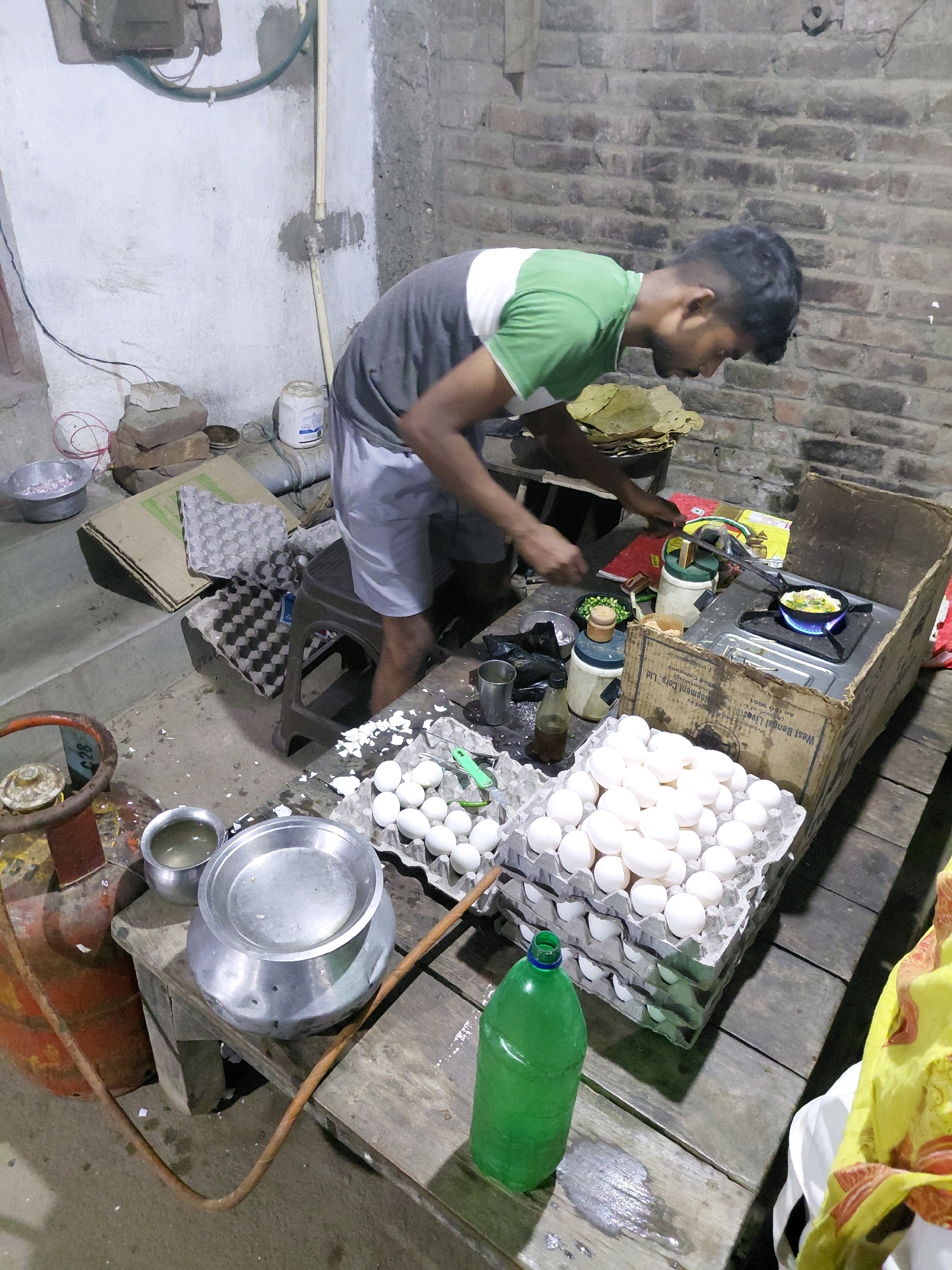 The image depicts a street food vendor preparing food in Patna, India. The scene is set outdoors, likely in a narrow alley or makeshift stall, with a brick wall forming the backdrop. A young man, wearing a grey and green t-shirt and light-colored shorts, is actively engaged in cooking. He is leaning over a small gas stove, where a pan with food is sizzling.
In front of him, several stacks of egg cartons are visible, filled with white eggs. A green plastic bottle, likely containing water or another beverage, sits on a wooden table. Adjacent to the eggs, there's a large metal pot and a smaller metal bowl containing liquid. A propane gas cylinder is also present, connected to the stove by a hose. Various other kitchen utensils and food items, including chopped green chilies and what appears to be a stack of plates made of dried leaves, are scattered around the cooking area. The overall environment suggests a basic and functional setup for preparing and selling food, typical of street vendors in this region.