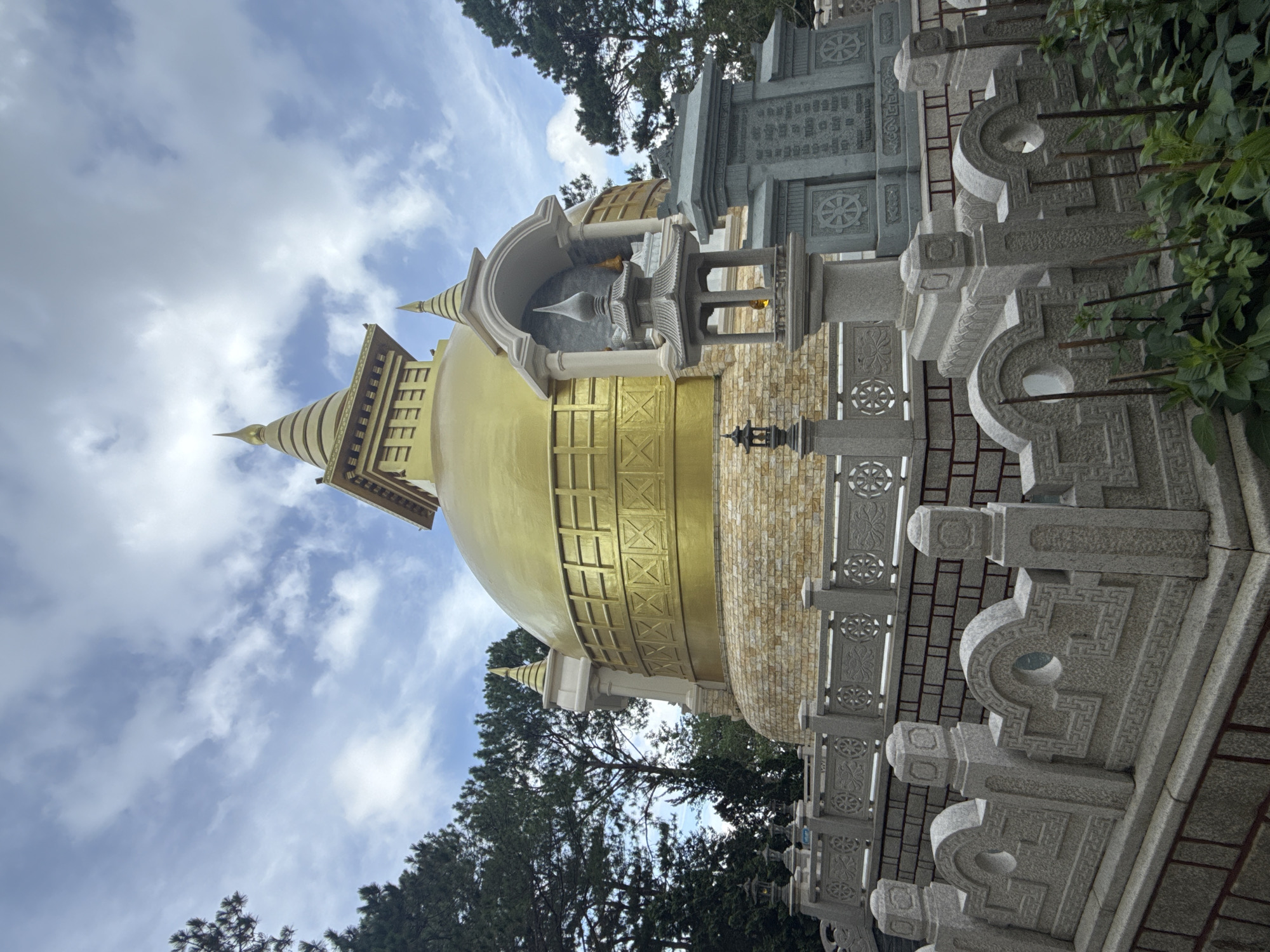 The image shows the Golden Pagoda (Truc Lam Zen Monastery) in Da Lat, Vietnam. The pagoda is a large, golden stupa with multiple tiered roofs and intricate detailing. It is surrounded by a stone courtyard with decorative railings and smaller structures. Trees are visible in the background, and the sky is partly cloudy. The scene is peaceful and serene, with no people visible. The pagoda is a prominent landmark in Da Lat and is a popular tourist destination.