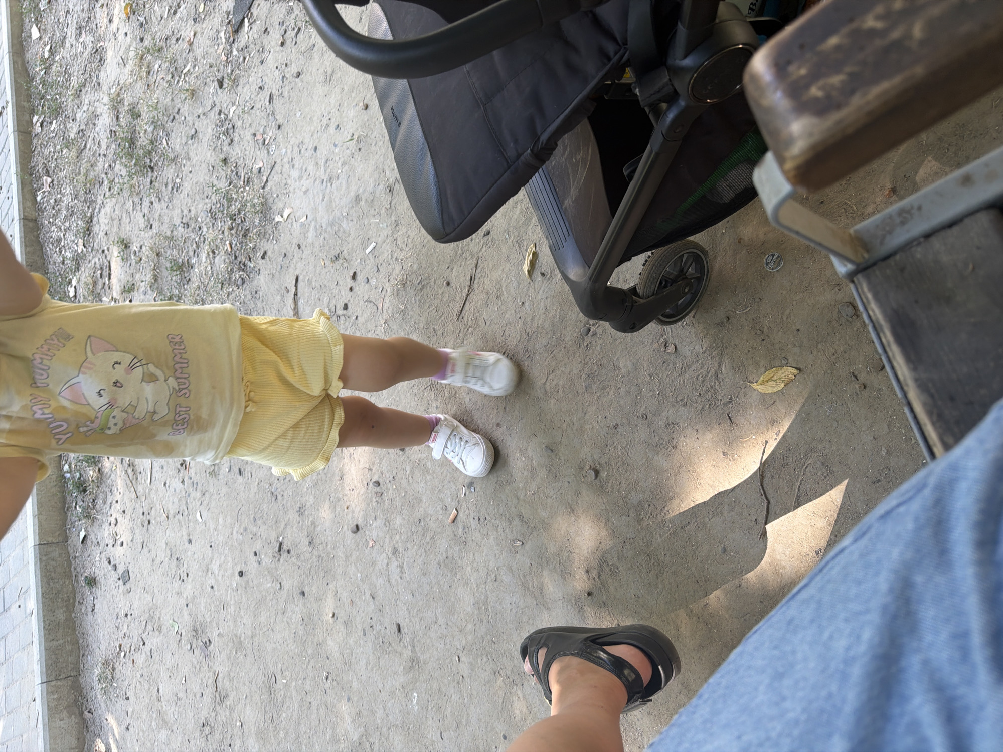 The image depicts a ground-level view of a park or outdoor recreational area. In the foreground, a person's legs clad in blue denim are visible, suggesting they are seated. To their left, another person's foot wearing a black sandal is positioned. A child, standing in yellow shorts and a light-colored t-shirt with a cartoon cat graphic, is also present. The child is wearing white sneakers with pink socks. A black stroller is situated to the right of the child. The ground is covered with dirt and scattered leaves. The background shows a paved walkway and some sparse vegetation. The scene appears to be in Chișinău, Moldova, based on the metadata.