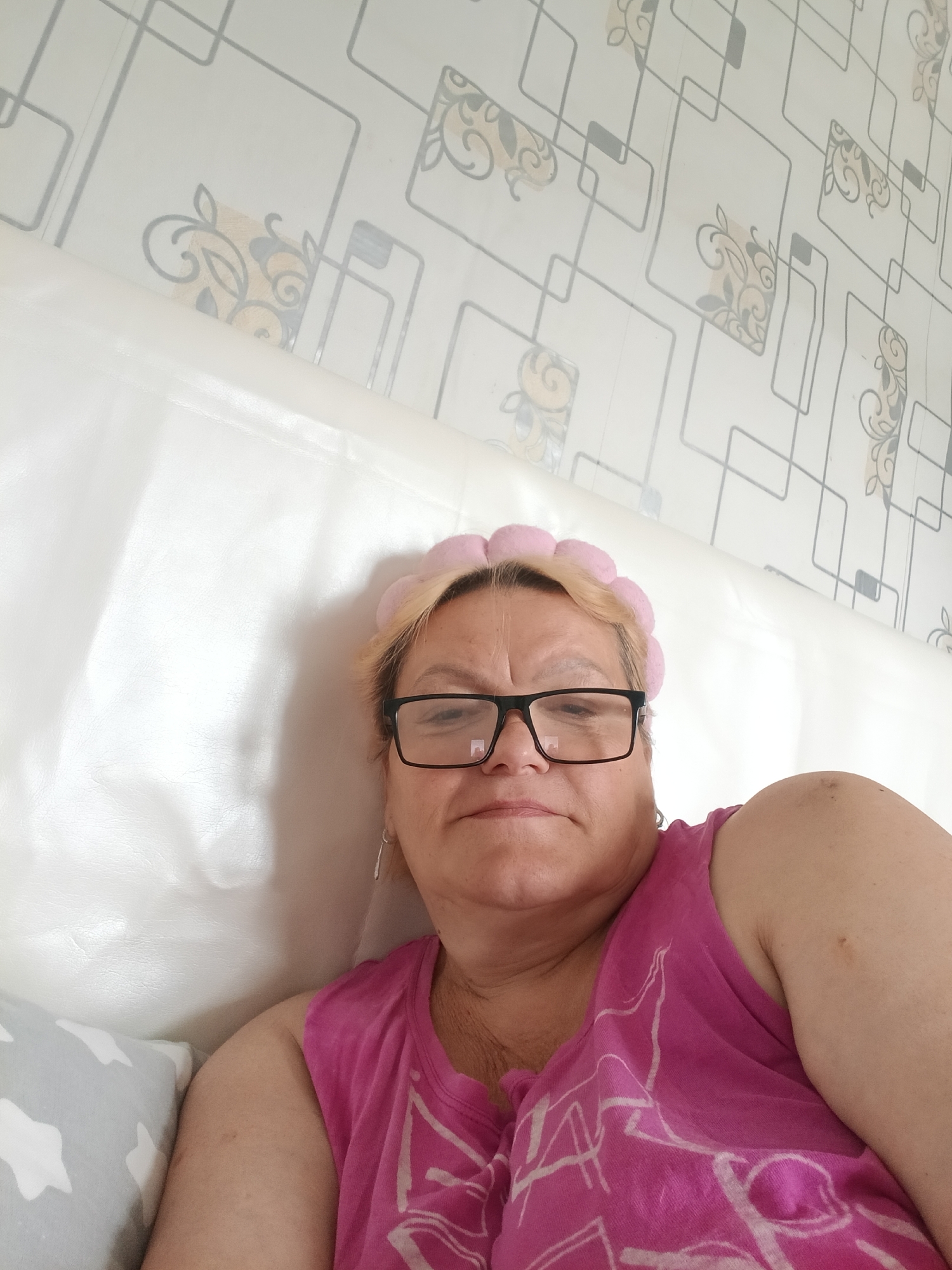 A medium, eye-level shot shows an adult female in Красный Маяк, Russia. She is wearing large, black-framed glasses and a pink shirt, and has a pink, padded headband. She appears to be reclining against a white headboard or cushion, with a light gray pillow at the lower left. The wallpaper behind the headboard features a geometric pattern of gray lines forming squares with beige floral designs. The woman is looking directly at the camera. She has fair skin, and her hair is blonde. A small silver earring is visible in her left ear.