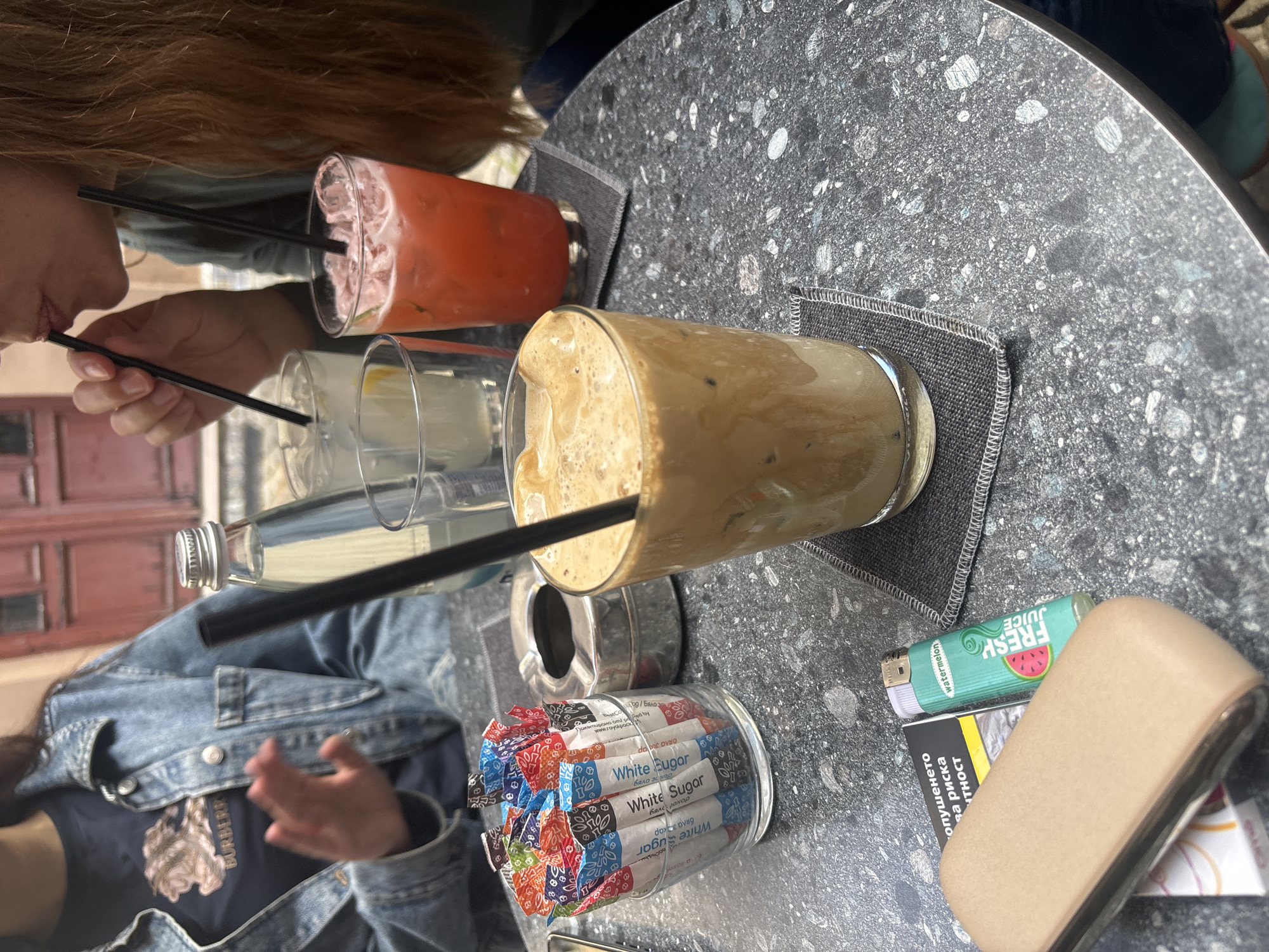 The image shows a close-up of a café table in Plovdiv, Bulgaria, with multiple drinks and personal items. Two individuals are partially visible, enjoying beverages. One person with long, reddish-brown hair is sipping a pink/red drink through a black straw. Another person, wearing a denim jacket over a dark t-shirt with a "BURBERR" logo, is also seated at the table.
On the dark, speckled table (possibly terrazzo), there are several drinks: a tall glass of iced coffee with a frothy top, the pink/red beverage, and a glass with a clear liquid and lemon slice, along with a glass bottle of water. All drinks feature black straws and rest on dark grey coasters.
Other items include a clear jar filled with colorful "White Sugar" packets, also labeled "бяла захар" in Cyrillic. An ashtray, a teal "watermelon JUICE FRESH" lighter, and a cigarette pack displaying "опушенето за риска" (Bulgarian for 'smoking for risk') are also on the table. The scene suggests a relaxed, social gathering in an outdoor setting during the daytime. The "Royalpack" text is visible on some sugar packets.