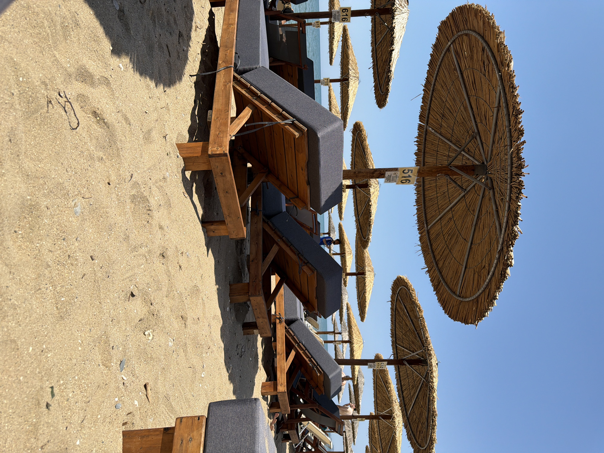 This image depicts a sunny beach scene in Alexandroupolis, Greece, viewed from a low angle. The primary subjects are rows of wooden sunbeds with dark grey cushions and large, circular thatched beach umbrellas. The setting is a sandy beach extending towards the clear blue sea, visible in the background.
The sand in the foreground is golden and appears fine, with some small pebbles and shells scattered across it. Strong, defined shadows cast by the sunbeds and umbrellas indicate bright daylight, likely midday or early afternoon, under a clear, cloudless blue sky.
Several sunbeds are visible in the foreground, neatly arranged. Further down the beach, multiple individuals can be seen enjoying the sun or relaxing on other sunbeds, though they are small and indistinct. One person, possibly a child, is faintly visible closer to the water's edge.
Numerous straw umbrellas line the beach, each mounted on a wooden pole. Some poles feature small white signs with black numbers. Visible numbers include "616", "516", and "55". Other signs display smaller numerical pairs like "7:5", "6:5", and "3:5", which might indicate pricing, zone identifiers, or other operational details. The overall scene conveys a typical summer vacation atmosphere, suggesting relaxation and leisure activities at the beach.