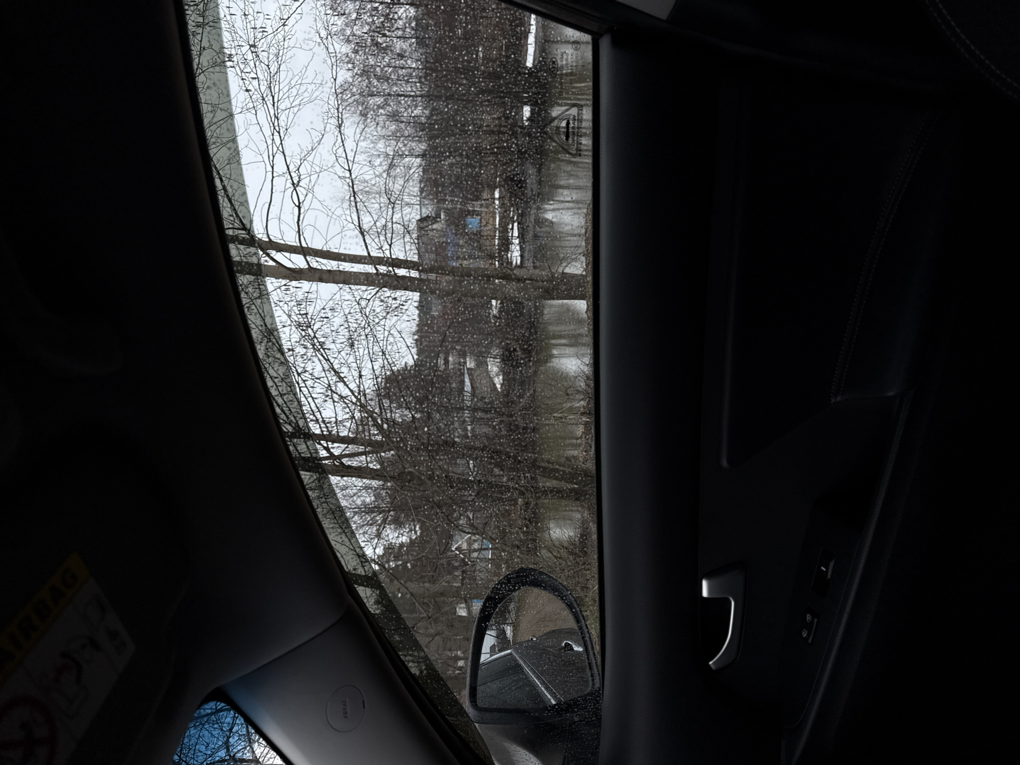 The image shows a view from inside a car on a rainy day. Through the car window, bare trees are visible, partially obscuring buildings in the background. Raindrops cover the window. A side mirror is also visible, reflecting another car. The car interior is dark, with the door panel and some control buttons visible. Based on the building style visible through the window, it's difficult to definitively determine the location but some buildings in Chaikovsky, Russia have a similar architectural style. Without further information or more distinct landmarks, a precise location within the city can't be confirmed.