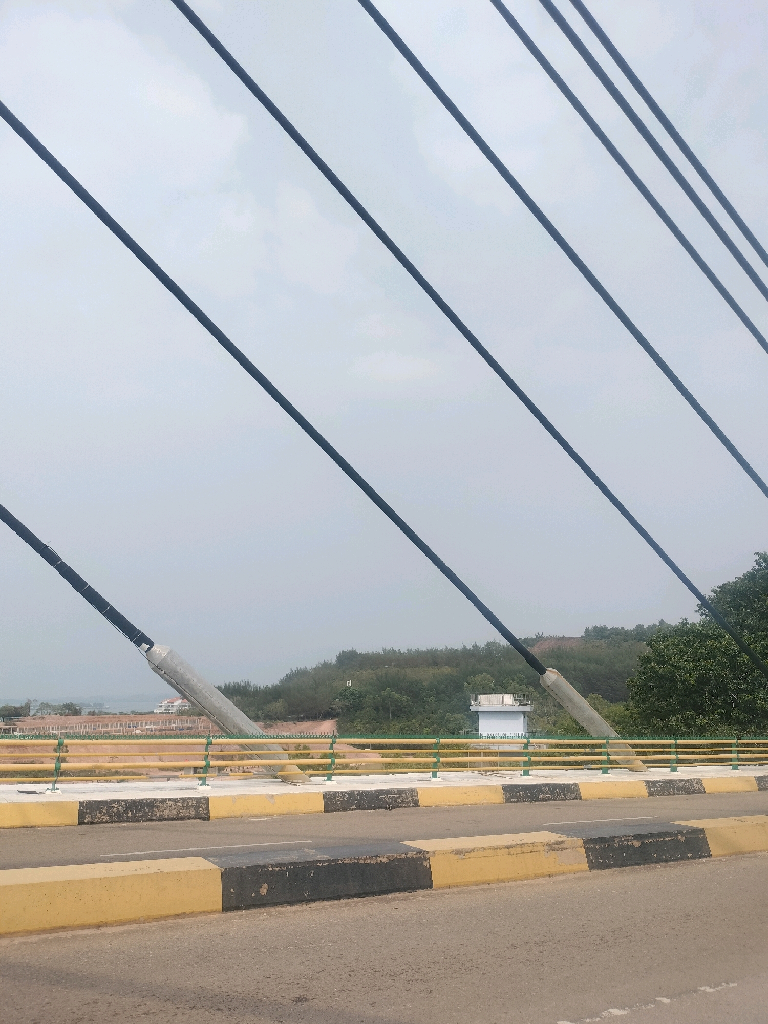 The image, captured from a vehicle on a bridge, displays a section of the Barelang Bridge in Batam, Indonesia, under an overcast sky. The foreground features a asphalt road with yellow and black painted concrete barriers. Beyond these barriers, the bridge's railing is visible, consisting of yellow horizontal bars supported by green posts. Diagonal black cables, characteristic of a suspension bridge, stretch across the frame towards the top. In the middle ground, a light blue building with a white cylindrical structure on top stands amidst a backdrop of green trees and rolling hills. The background reveals a hazy landscape with distant structures and vegetation. No people are present in the frame.