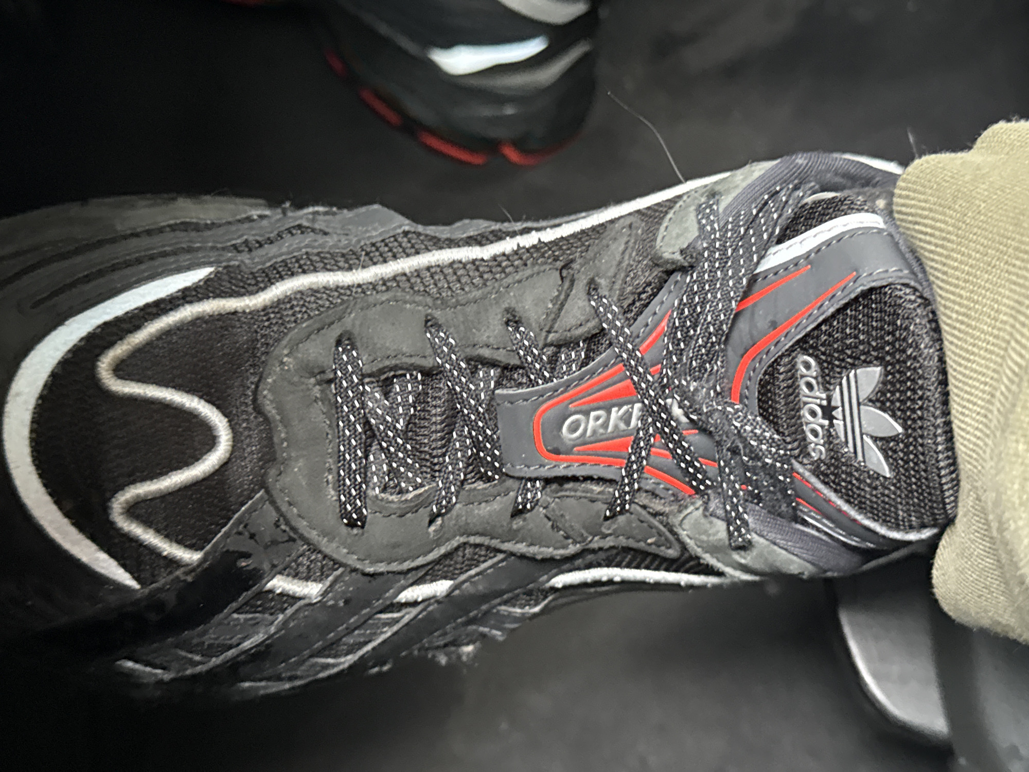 A close-up shot shows an Adidas Orketro sneaker on a person's foot. The shoe is predominantly black with white and red accents. The shoelaces are black with white speckles. The "Orketro" brand name is visible on the side of the shoe, and the Adidas logo is on the tongue. The setting appears to be indoors. There is not enough information to determine the location as Stara Zagora, Bulgaria.