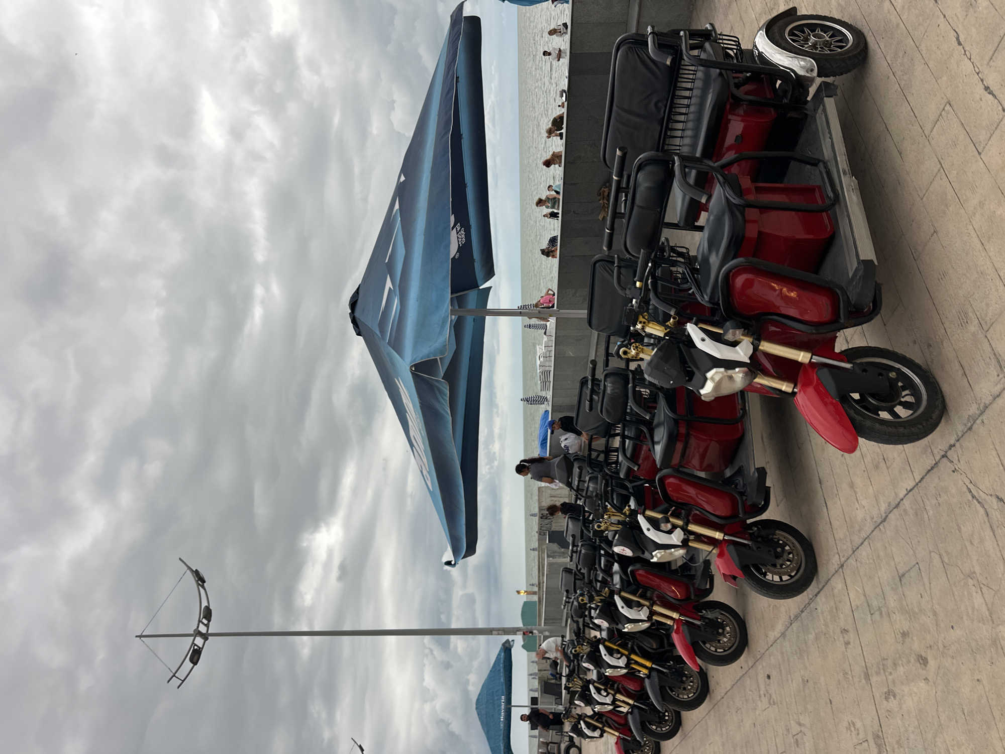 The image depicts a row of parked red and black electric scooters on a paved walkway, likely in Batumi, Georgia. In the background, beyond a low wall, is the sea with several people visible enjoying the water. The sky is overcast with thick, grey clouds, suggesting cloudy weather.
The scooters are arranged in a line, facing away from the viewer. They appear to be designed for multiple passengers, with some models featuring two seats and others potentially more. The scooters are a mix of red and black, with silver accents on some of the wheels and handlebars. One of the scooters at the front has a white front fender.
In the middle ground, a large, dark blue umbrella with white patterns is partially obscuring the view of the sea and the people. The umbrella has text visible on it, though it is not entirely legible. The people in the sea are scattered, some appearing to be wading or swimming.
Further in the background, a structure that looks like a promenade or boardwalk runs along the coastline. A tall lamppost with multiple lights is visible on the left side of the image. The overall impression is of a seaside town on a cloudy day, with recreational activities taking place by the water and transportation options readily available. The image is taken at an angle, making the scene appear somewhat tilted.