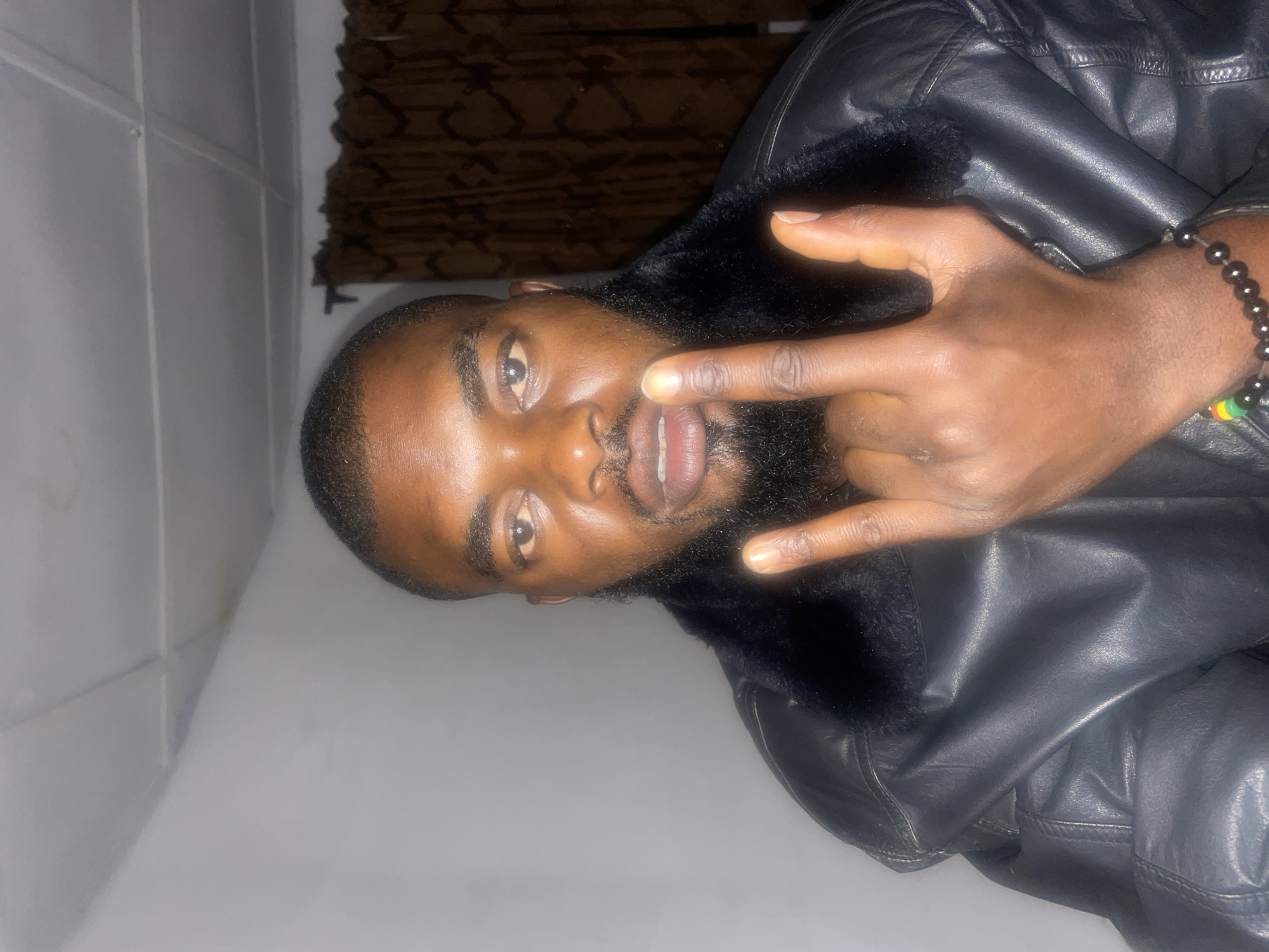 A medium shot depicts a man, likely in his 30s, wearing a black leather jacket with a fur collar. The man has dark skin, short black hair, and a black beard. He is making a hand gesture, resembling the "rock on" sign, near his face. The setting appears to be indoors, with a light-colored wall behind him and a dark brown patterned fabric in the background, possibly a curtain. The image does not contain sufficient information to definitively state that it was taken in Jos, Nigeria.